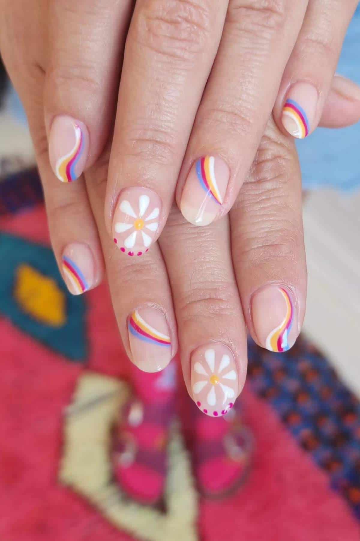 Close-up of hands with manicured nails featuring white daisy flower designs and colorful curved stripes on a pink and blue background.