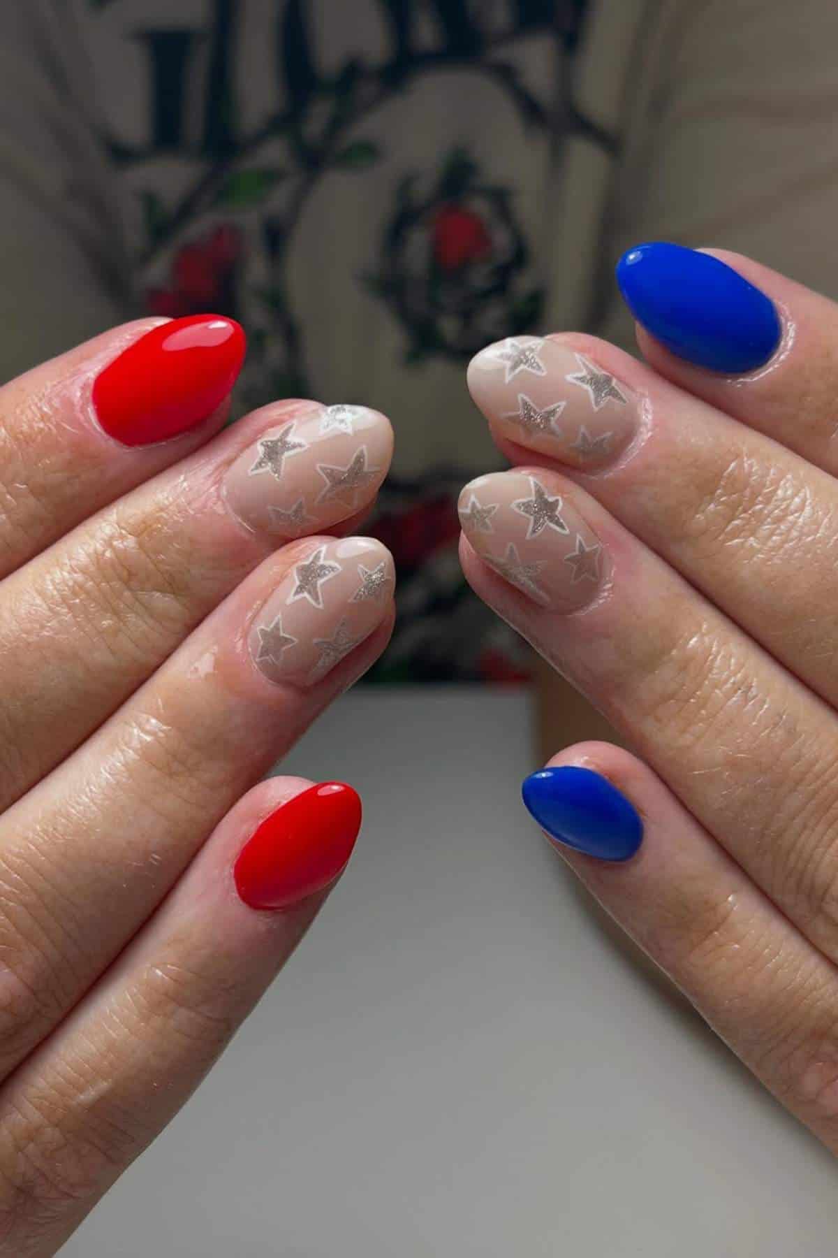 Close-up of hands with manicured nails featuring red, blue, and nude polish with silver star designs on the nude nails.