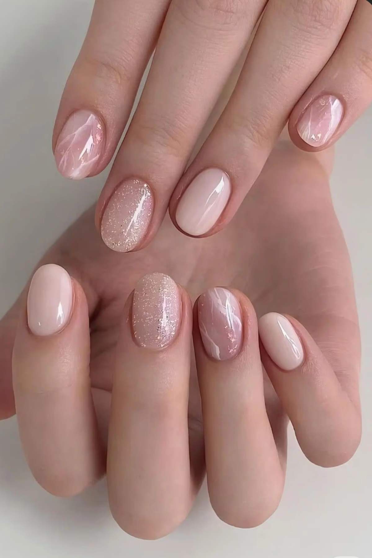 A hand with short, oval-shaped nails painted in light pink tones, featuring glossy, shimmery, and marbled designs.
