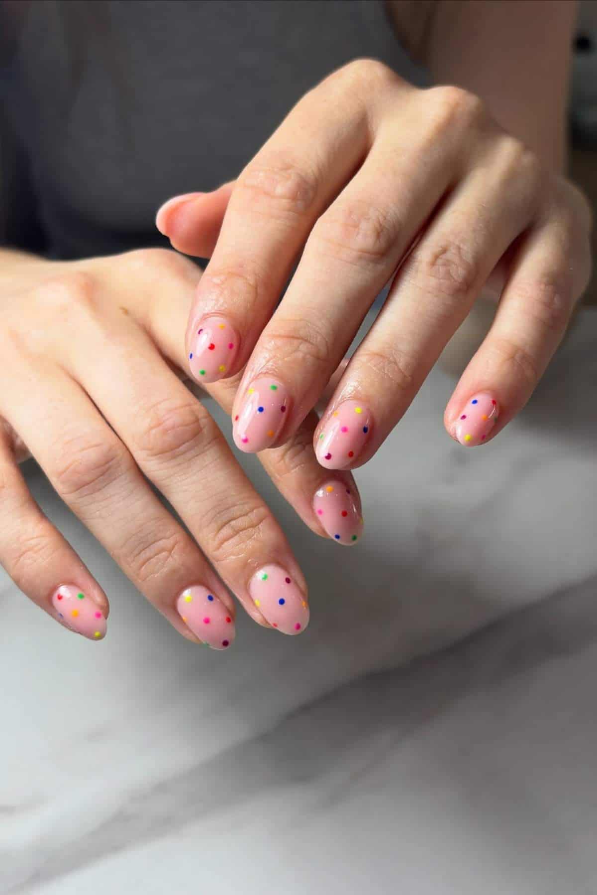 Close-up of hands with short, almond-shaped nails painted pink and decorated with multicolored polka dots, resting on a marble surface.