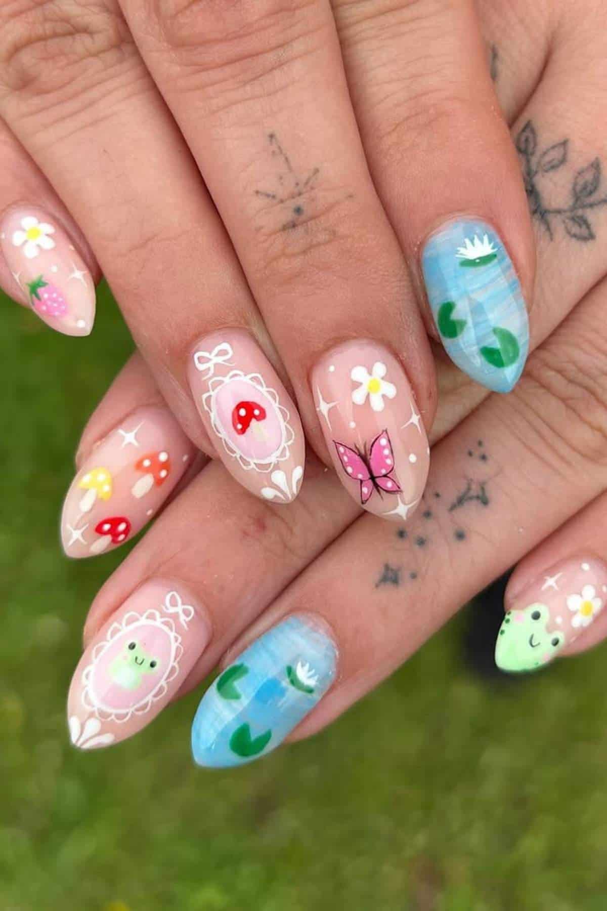 Hands with almond-shaped nails featuring colorful designs: strawberries, mushrooms, flowers, butterflies, lily pads, and smiling faces. Subtle hand tattoos are visible in the background.