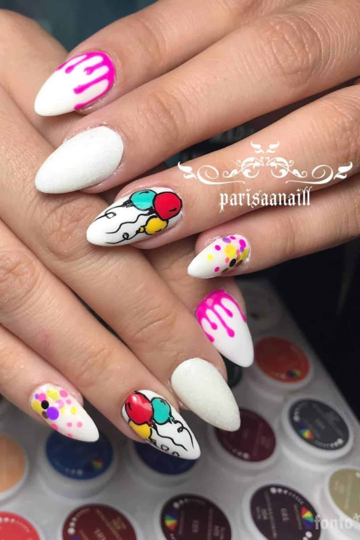 Close-up of hands with almond-shaped nails featuring white polish, colorful balloon nail art, pink drips, and multicolored dot designs.