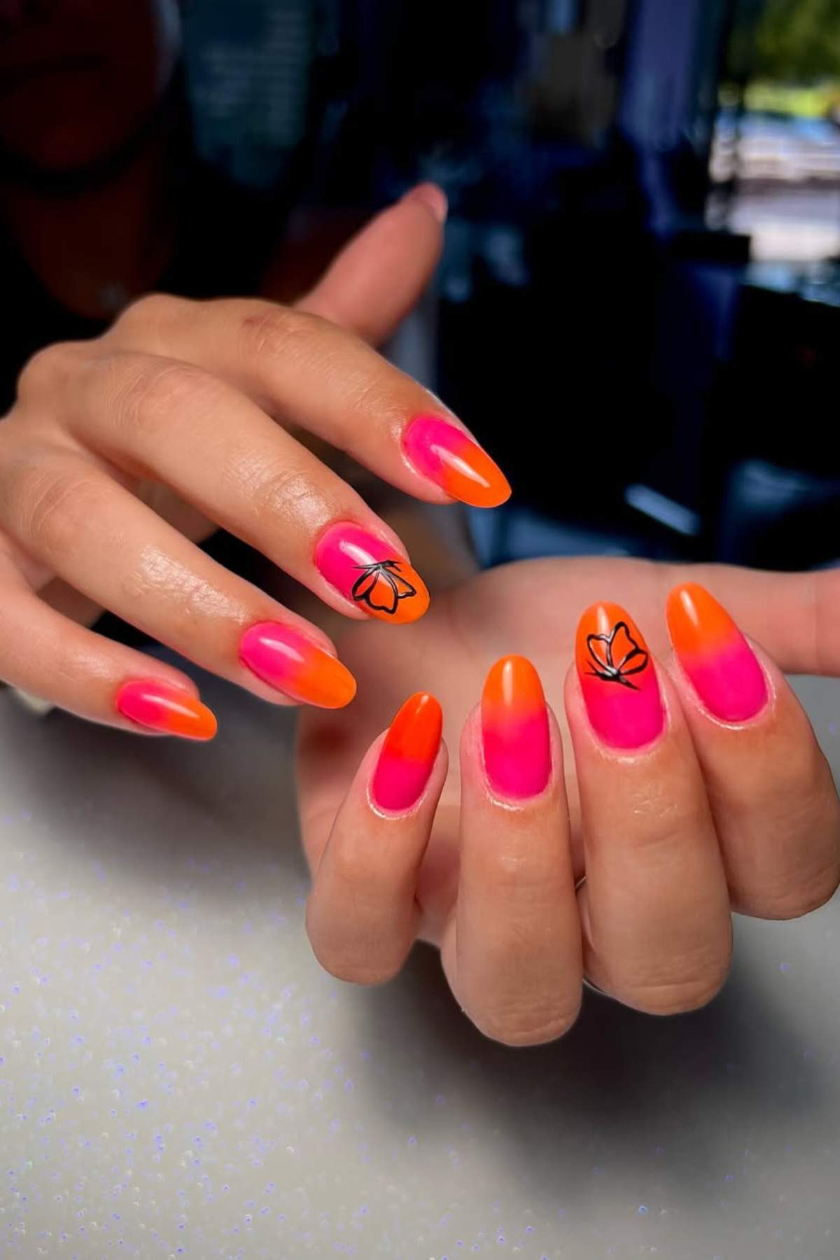 A hand with long almond-shaped nails painted in orange and pink ombre with black leaf designs on two accent nails.