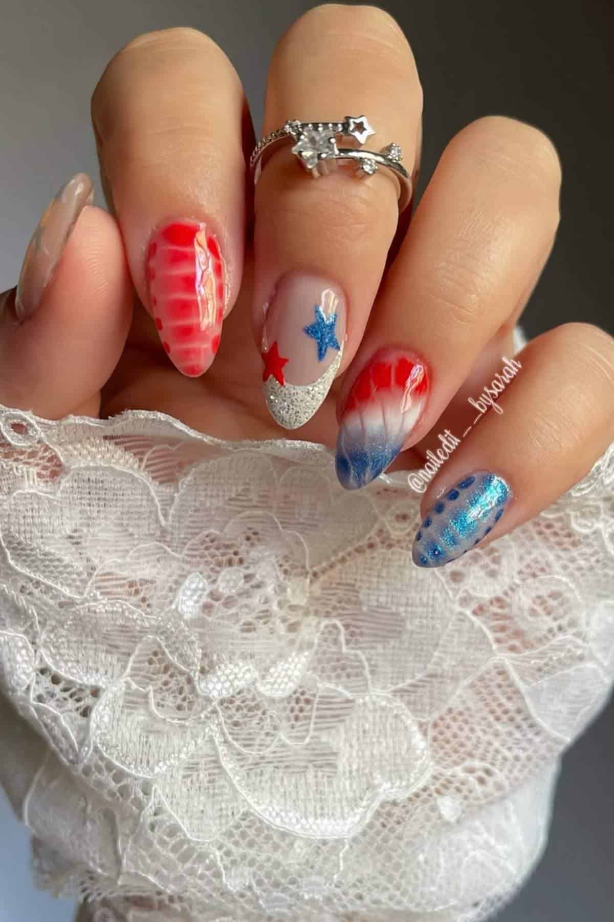 A hand with long nails painted in red, white, and blue designs, including stars and ombre effects, holds white lace fabric.
