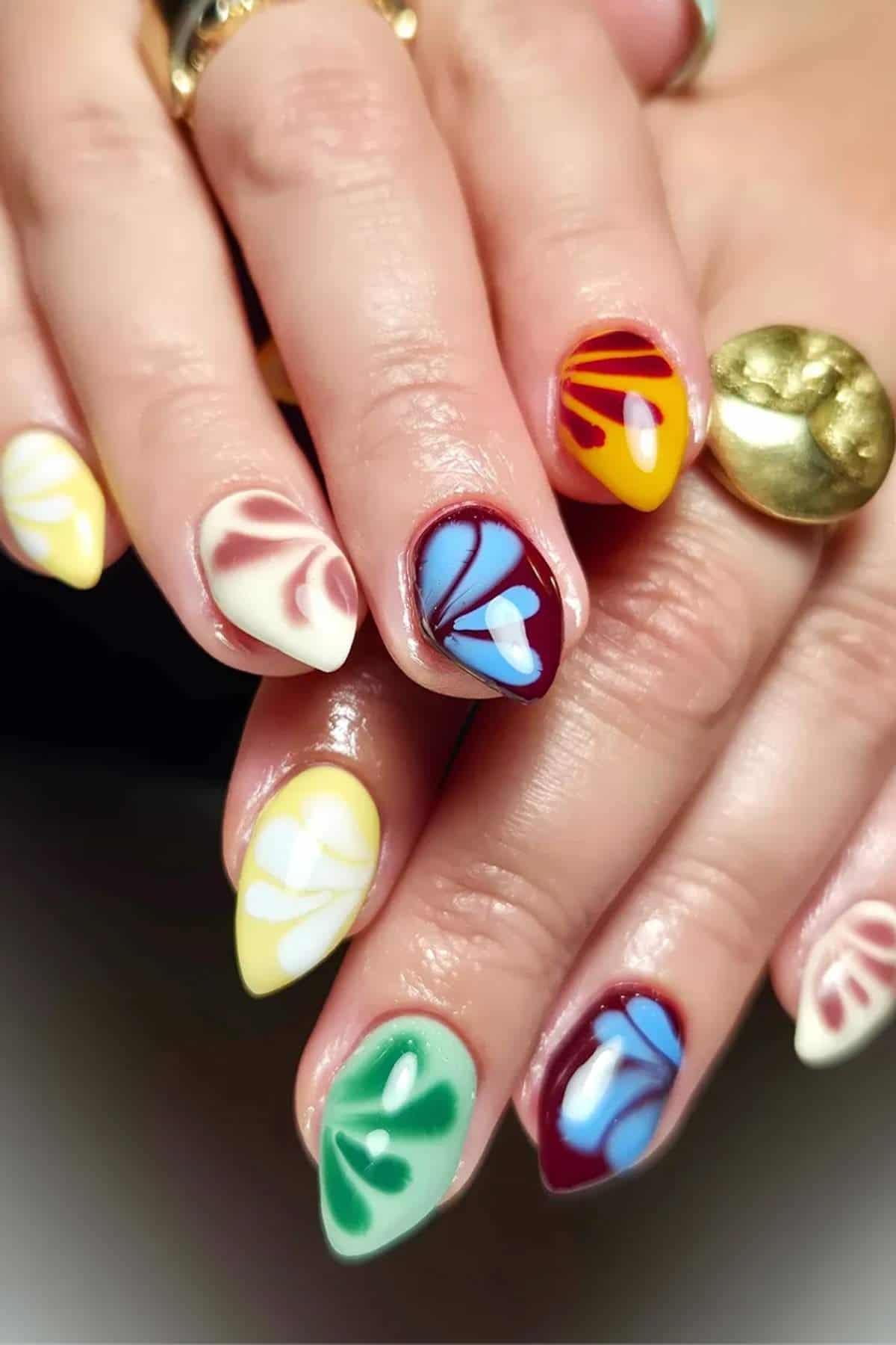 A close-up of hands with almond-shaped nails, each painted with colorful abstract floral designs in red, yellow, green, blue, and white.