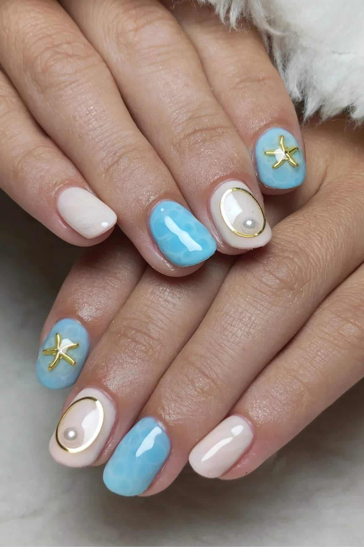 Close-up of hands with manicured nails featuring blue and white polish, gold starfish charms, pearl accents, and glossy finish.