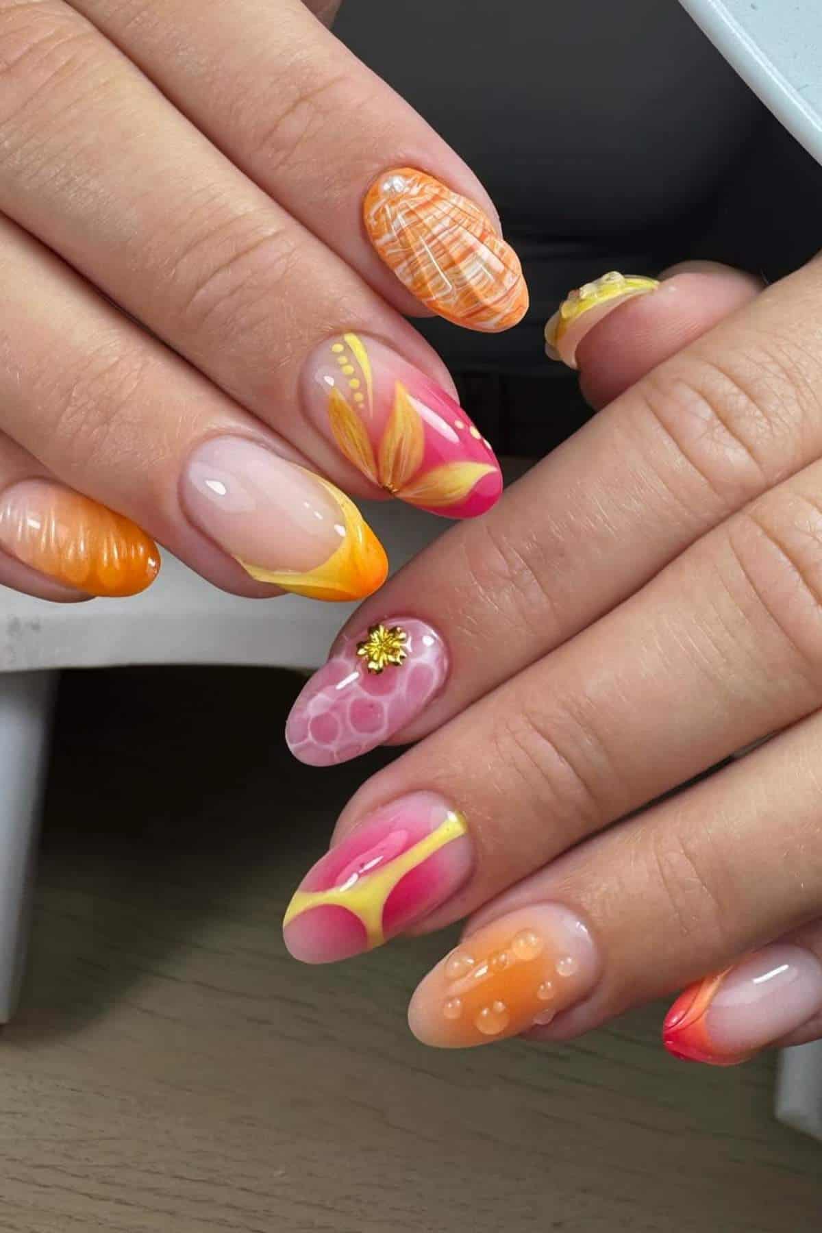 Hands with long, almond-shaped nails featuring various colorful designs in orange, yellow, and pink tones, including abstract patterns, flowers, and textured effects.
