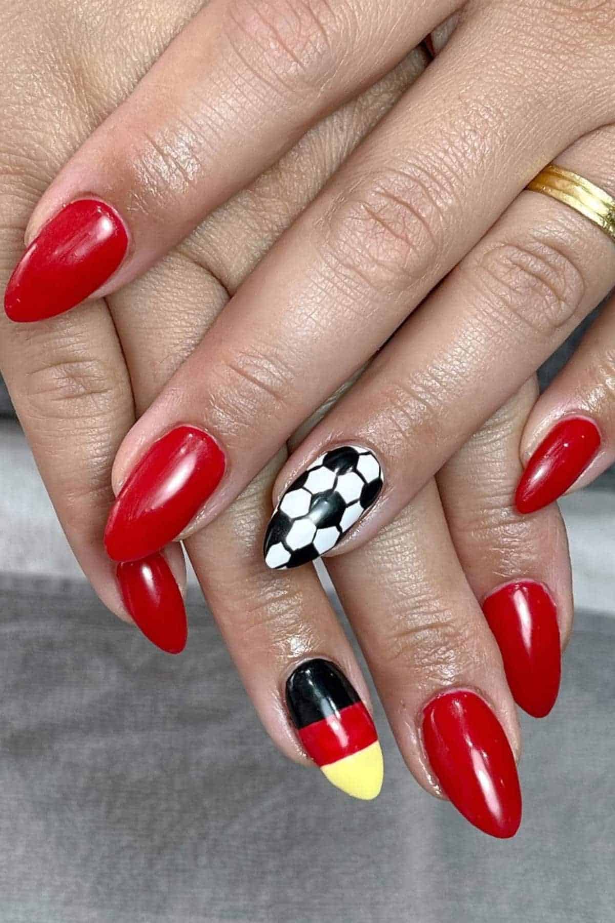 A hand with red nail polish; one nail has a soccer ball design, and another has a German flag design.