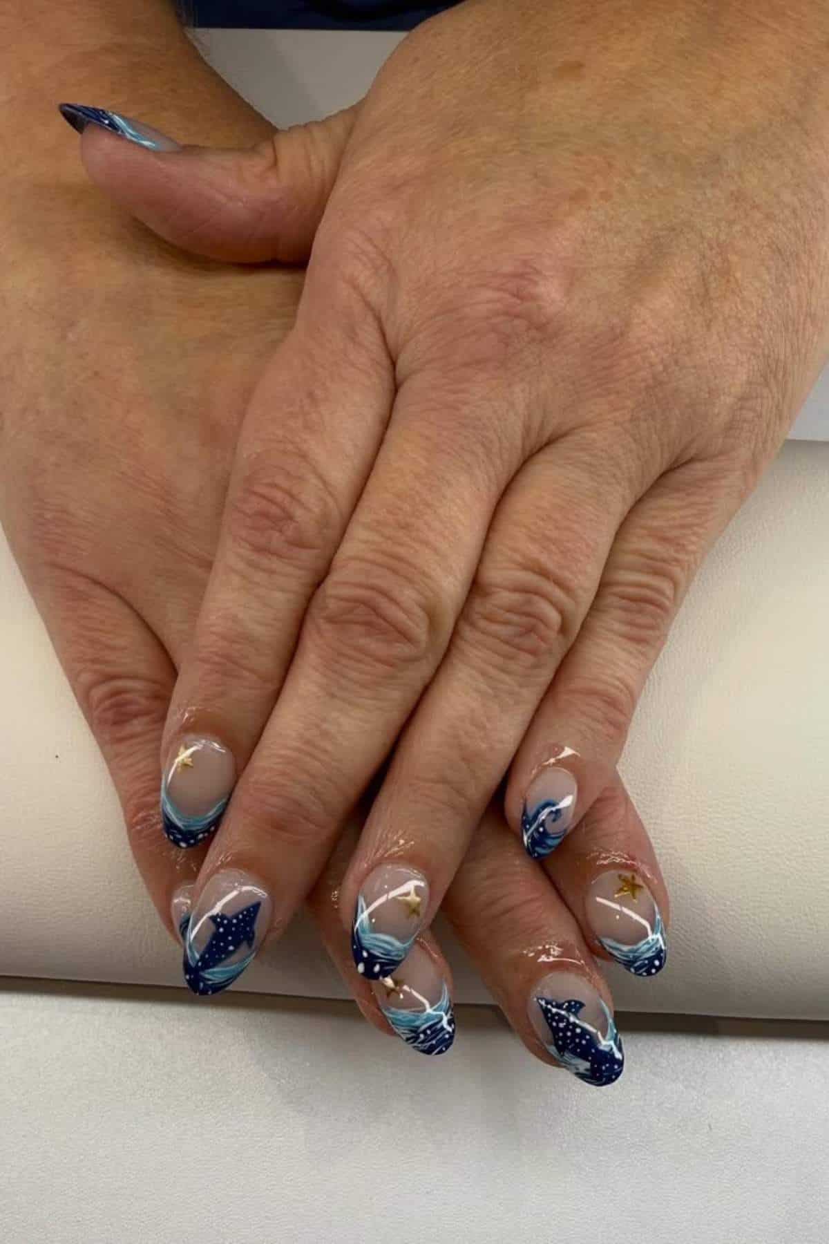 Hands with almond-shaped nails featuring blue and white wave designs, small star accents, and clear sections, resting on a light surface.