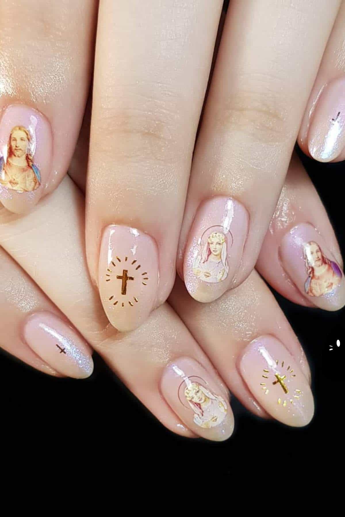 A close-up of hands with manicured nails, featuring designs of religious figures and gold crosses on a pale pink base.