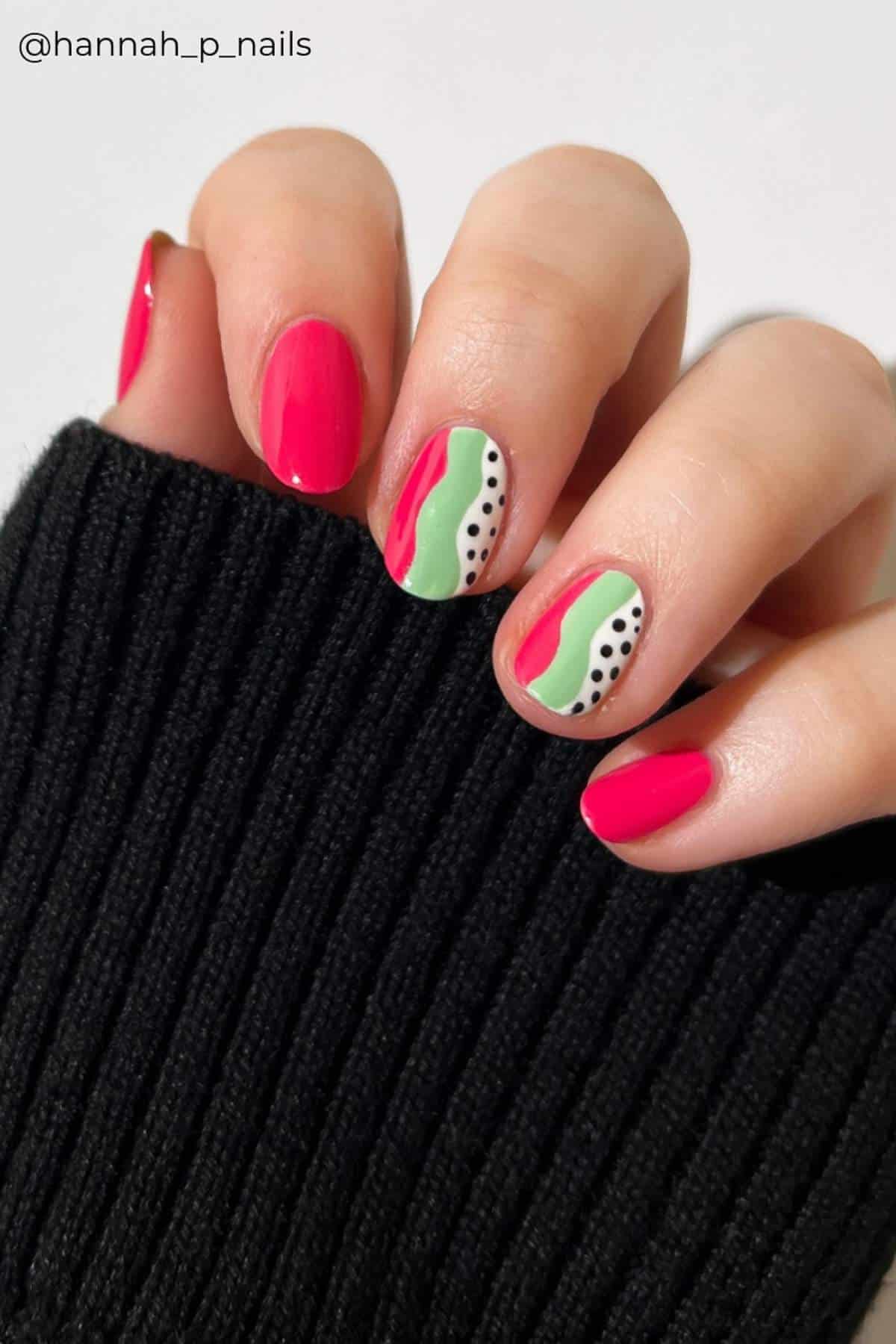 A hand with pink and accent nails; accent nails feature wavy green lines and black dots on a white background. The person is wearing a black ribbed sleeve.