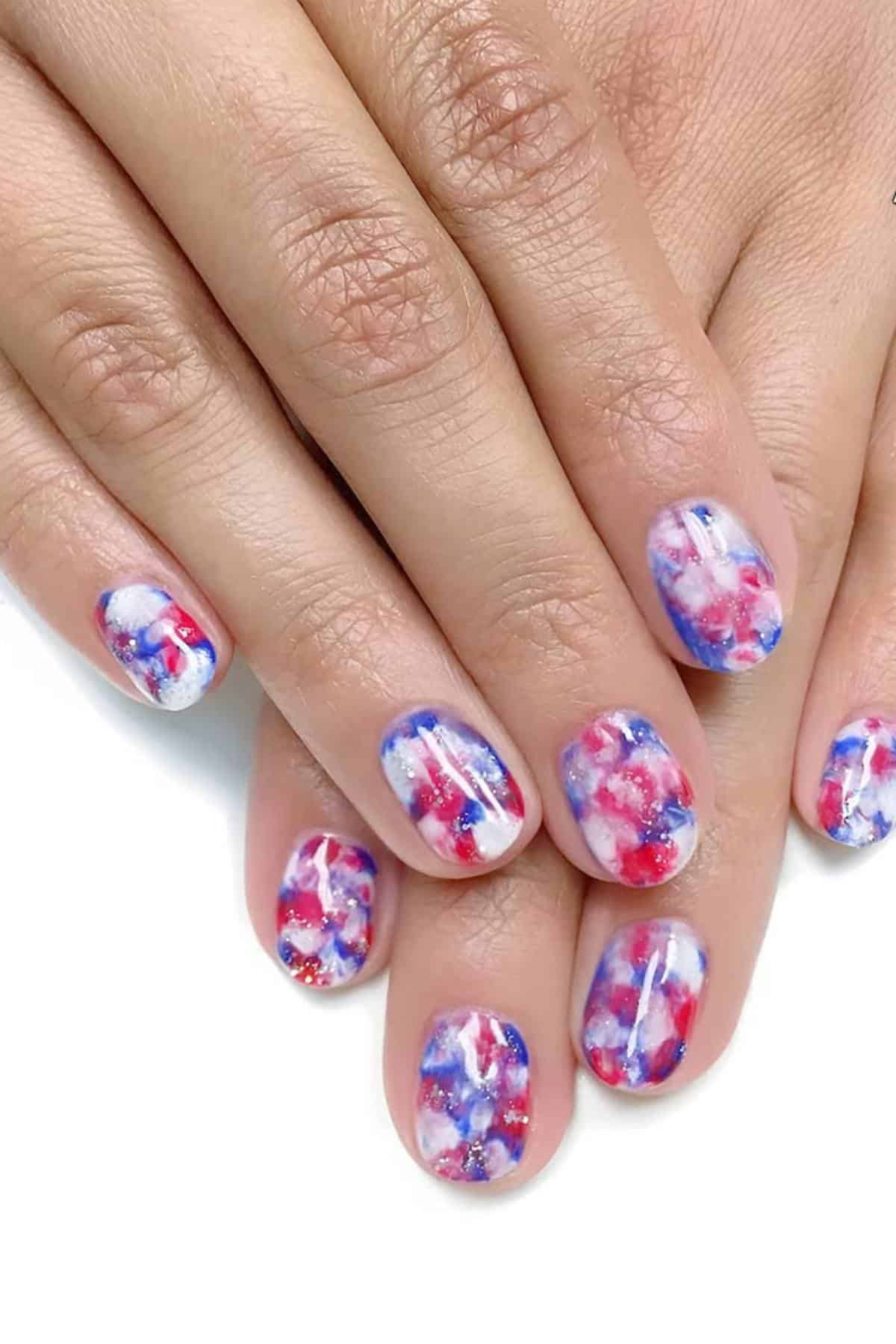 Hands with short fingernails featuring a red, white, and blue abstract marble nail design resting on a white surface.