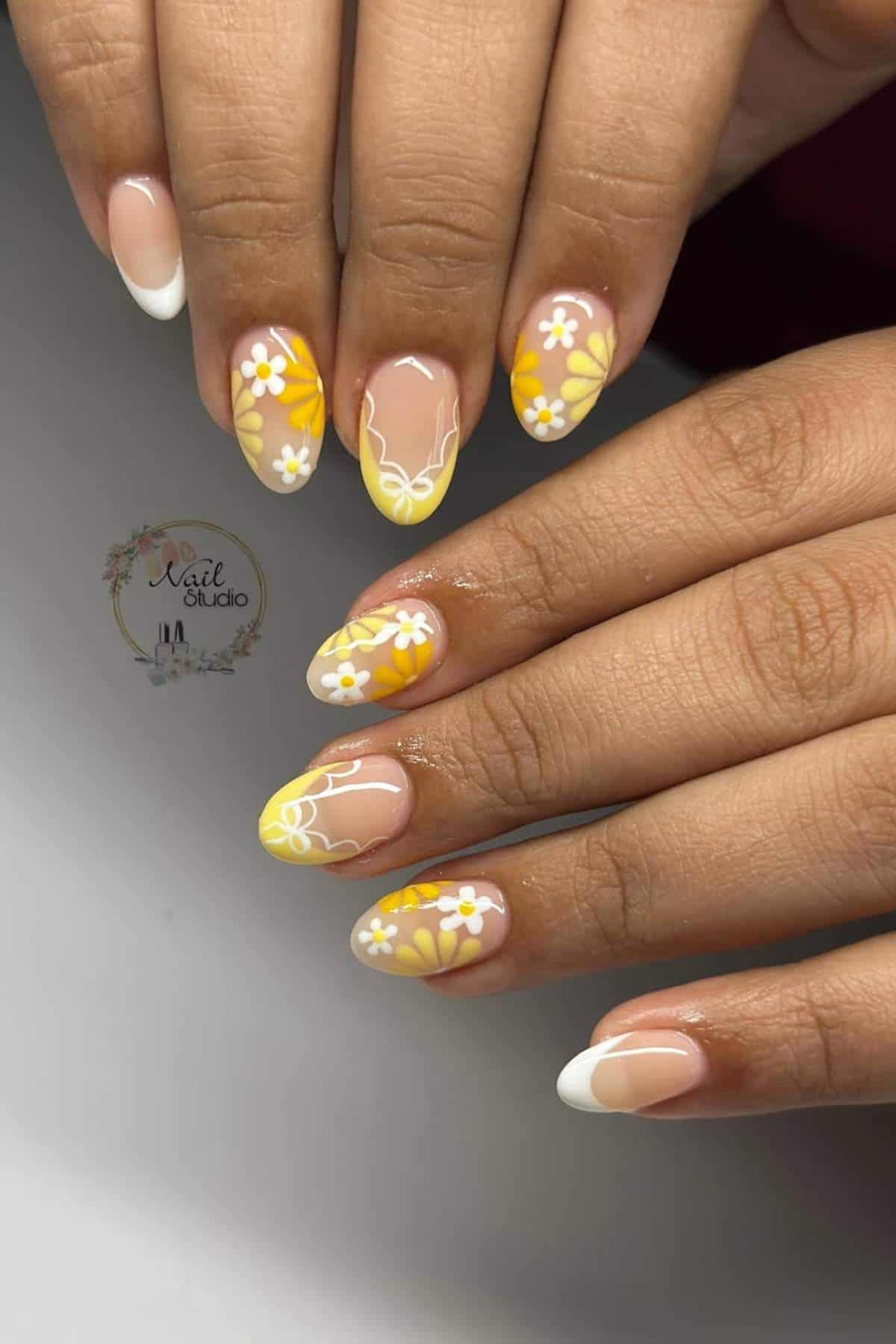 Hands with almond-shaped nails featuring a yellow and white floral design, accent nails with daisies, and classic French tips on some fingers.