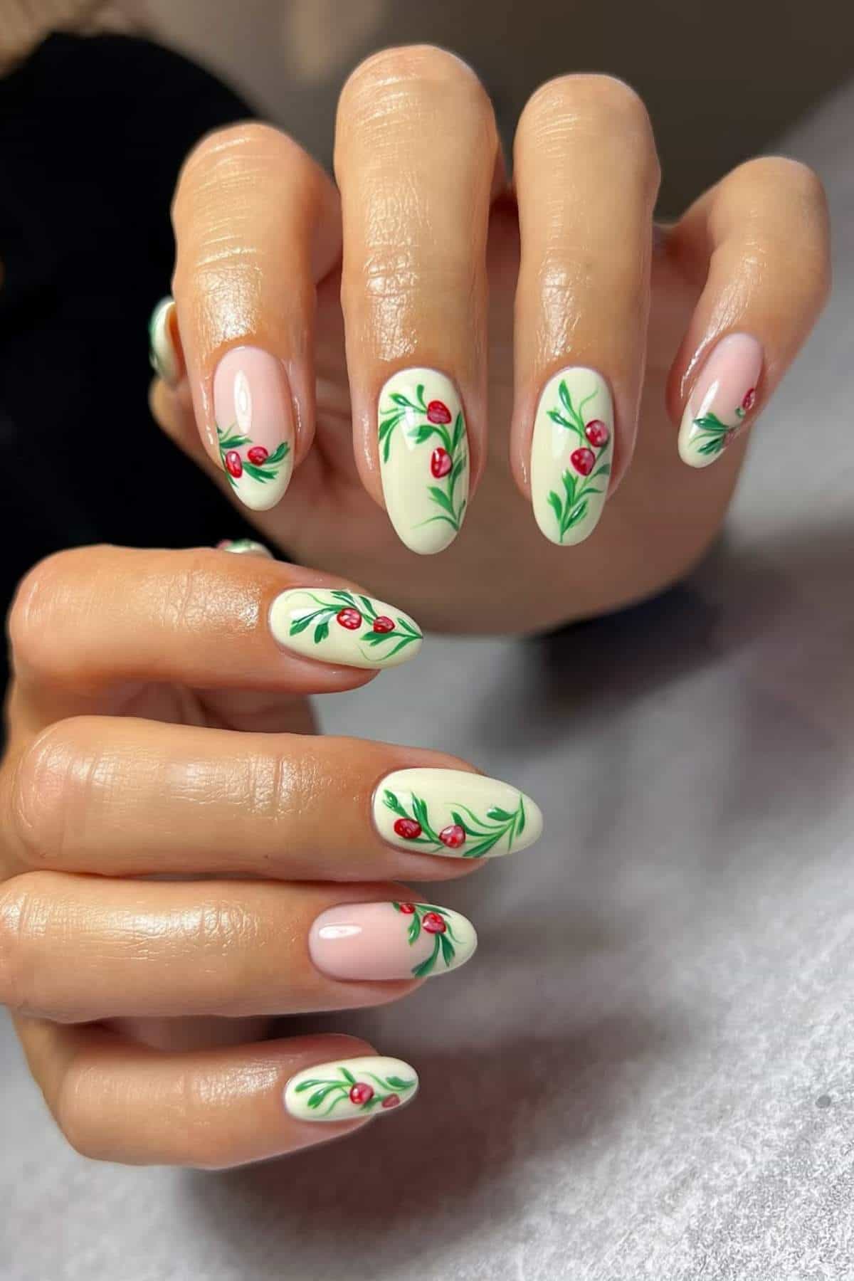 Close-up of hands with almond-shaped nails painted in light colors and decorated with green leaves and small red flowers.