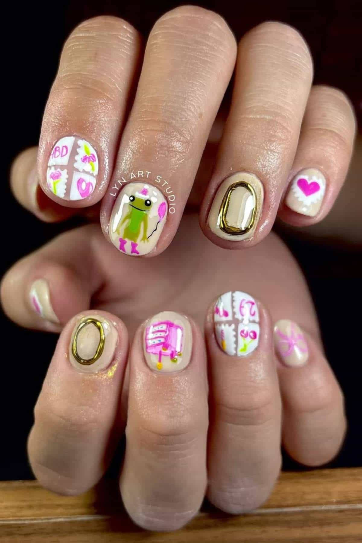 A close-up of fingernails with colorful nail art, including letters spelling "LOVE," a frog, flowers, hearts, and various small designs on a pale background.