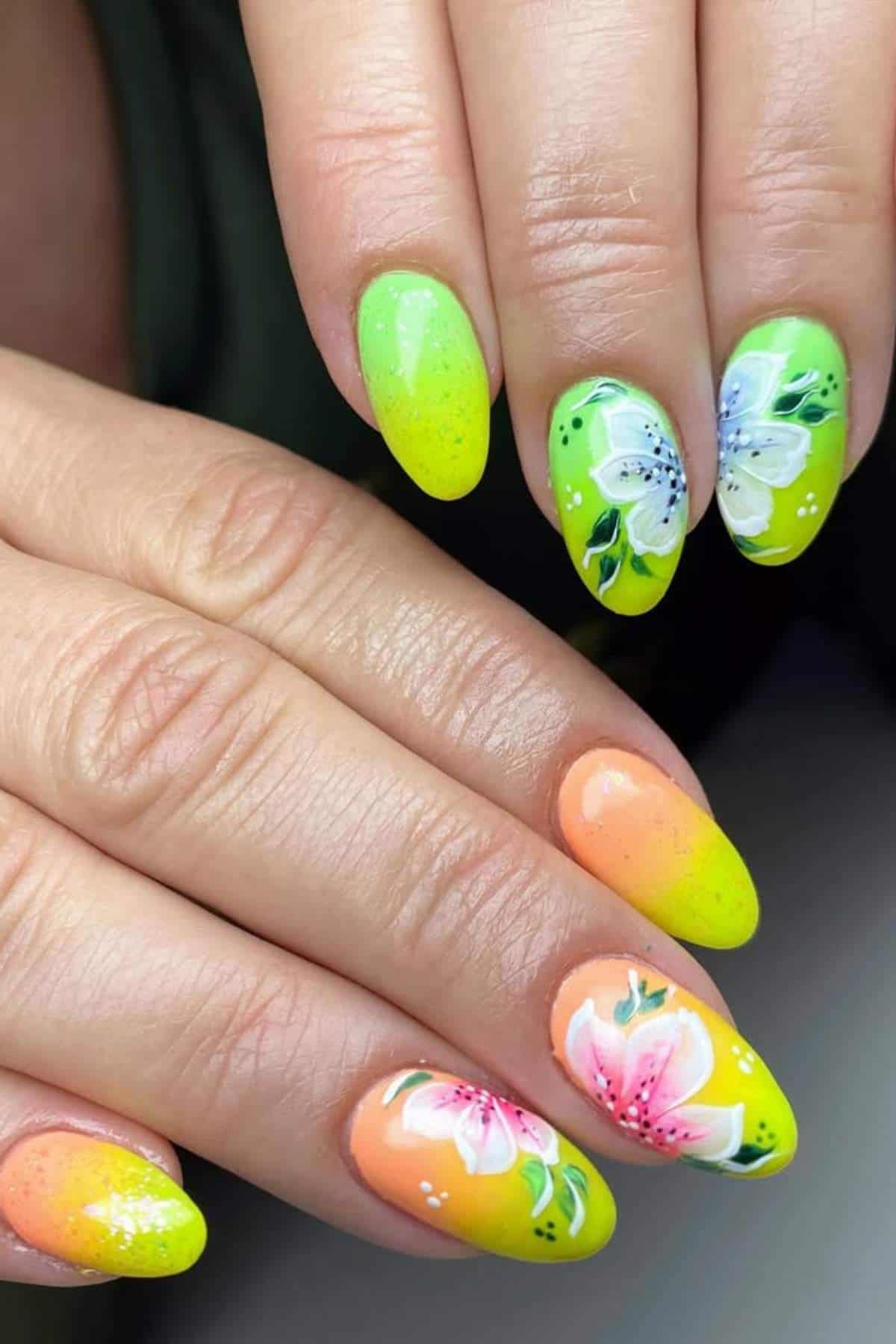 Hands with almond-shaped nails; one hand has green nails with white floral designs, and the other has orange nails with pink floral designs.