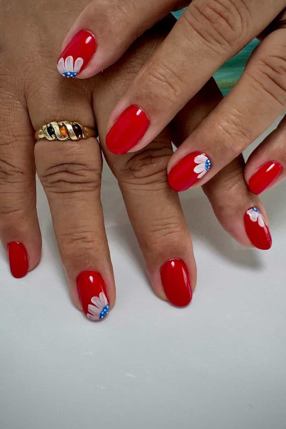 Close-up of hands with glossy red nail polish; some nails feature white daisy designs with blue accents near the cuticles. A gold ring is worn on one finger.
