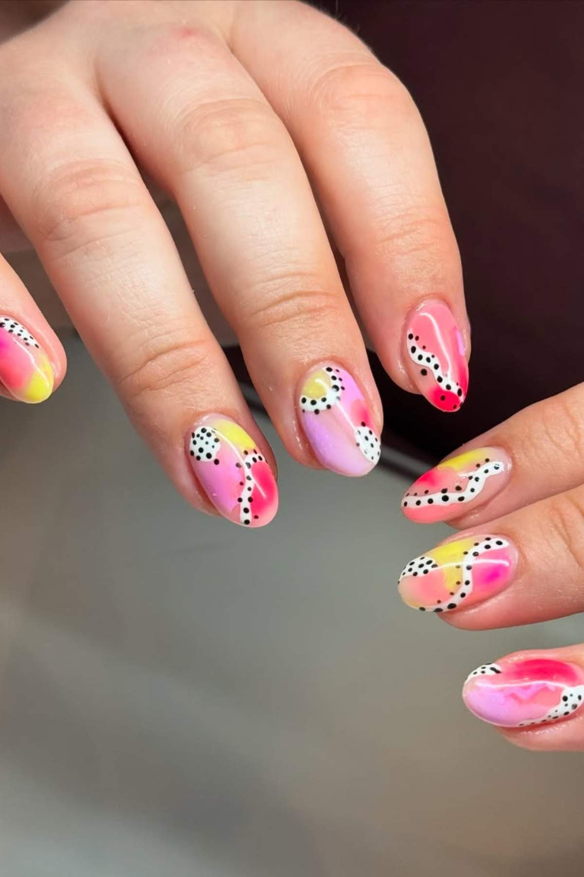 Hands with almond-shaped nails featuring abstract pink, yellow, and white designs, accented with black and white dotted lines.