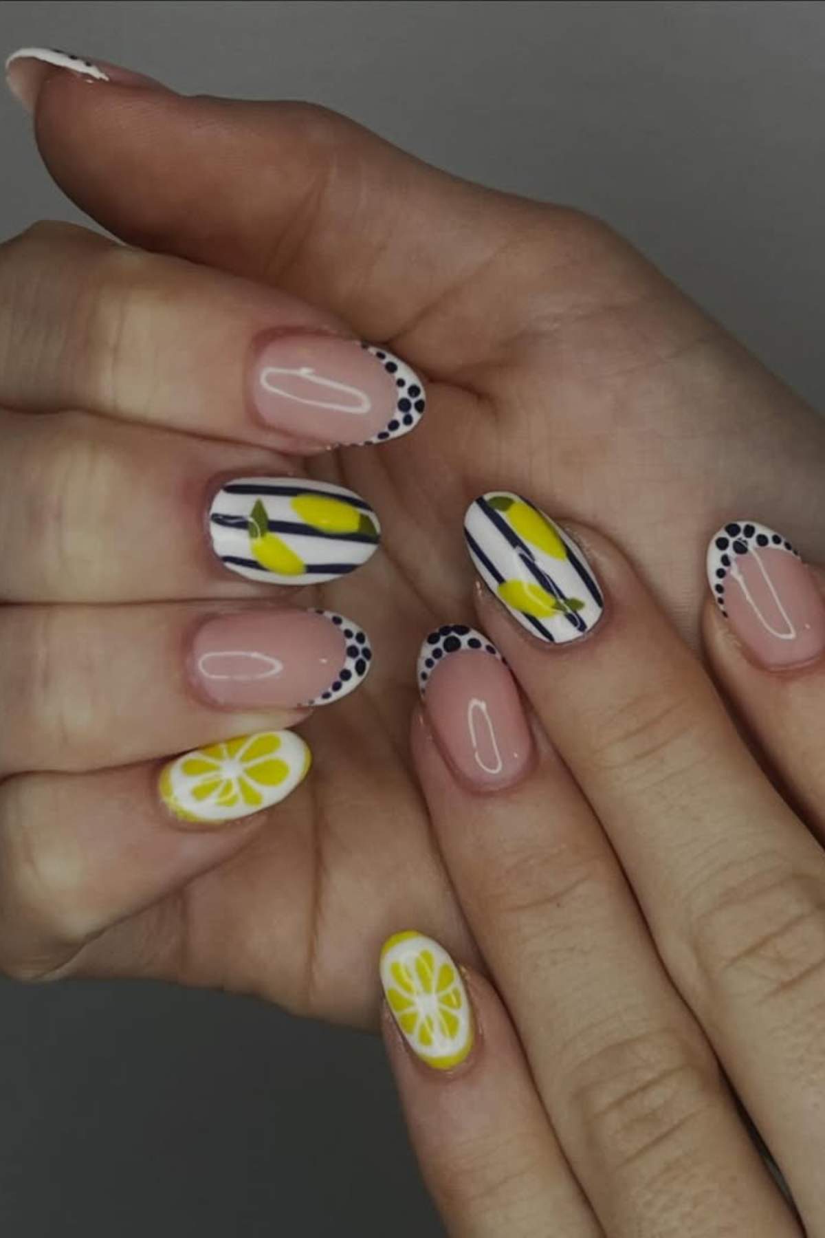 Hands with almond-shaped nails featuring a manicure with yellow and white lemon designs, black dots on the tips, and blue stripes on a few nails.