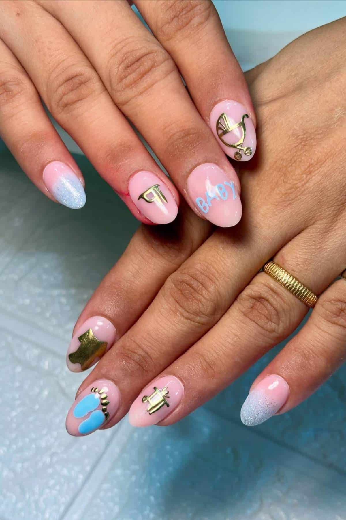 Close-up of hands with almond-shaped nails featuring a baby shower theme, including bottle, pacifier, rattle, stork, and "baby" nail art in blue, gold, and pink colors.