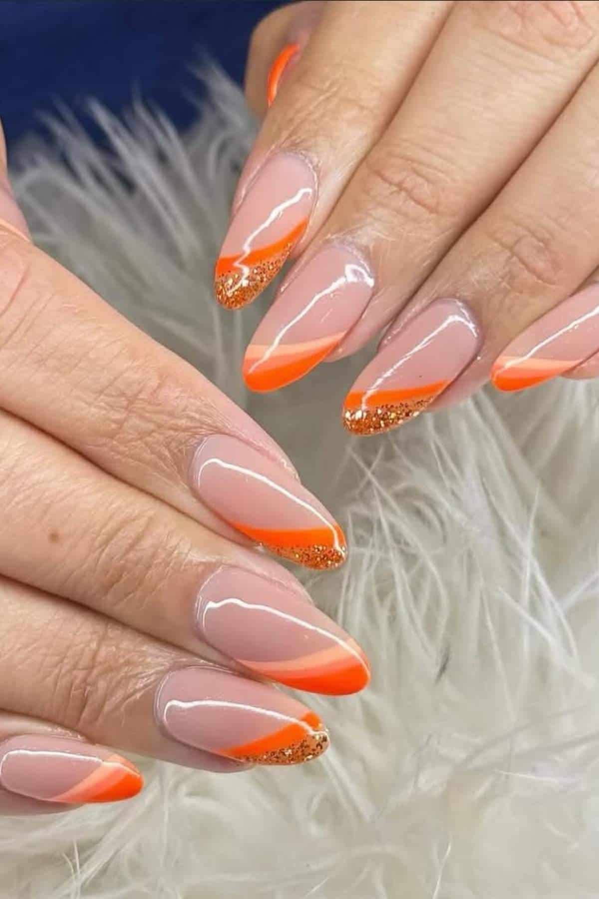 Hands with almond-shaped nails featuring a nude base, orange diagonal French tips, and gold glitter accents, resting on a white, fluffy surface.