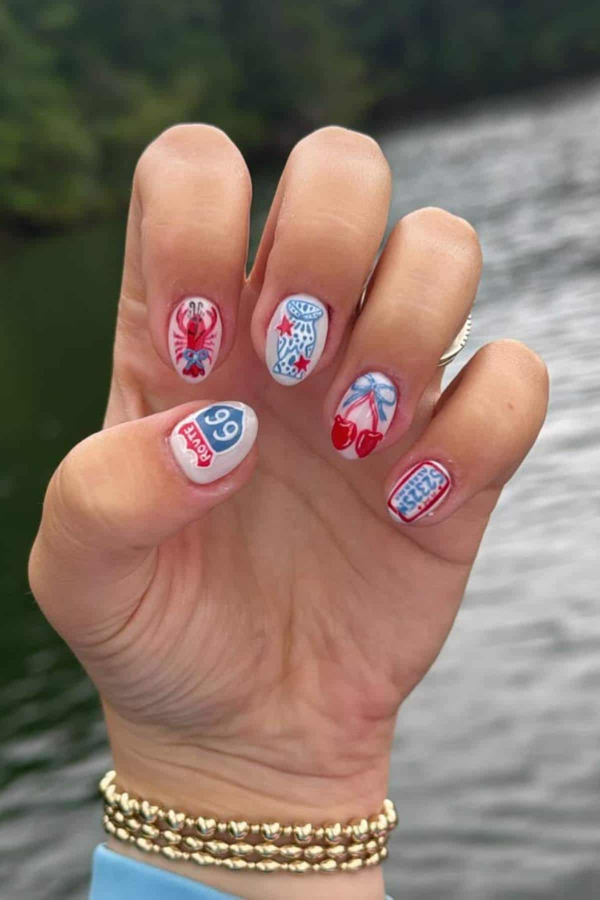 A hand with manicured nails featuring detailed red and blue designs, including a lobster, cherries, numbers, and patterns, held in front of a lake background.