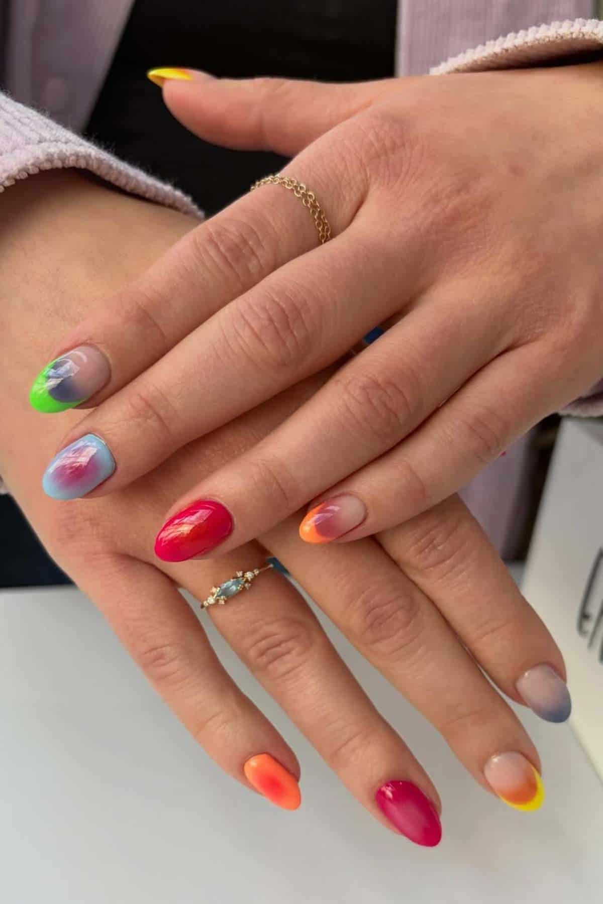 Close-up of hands with almond-shaped nails, each painted in different bright colors and gradient designs, with two rings on the fingers.