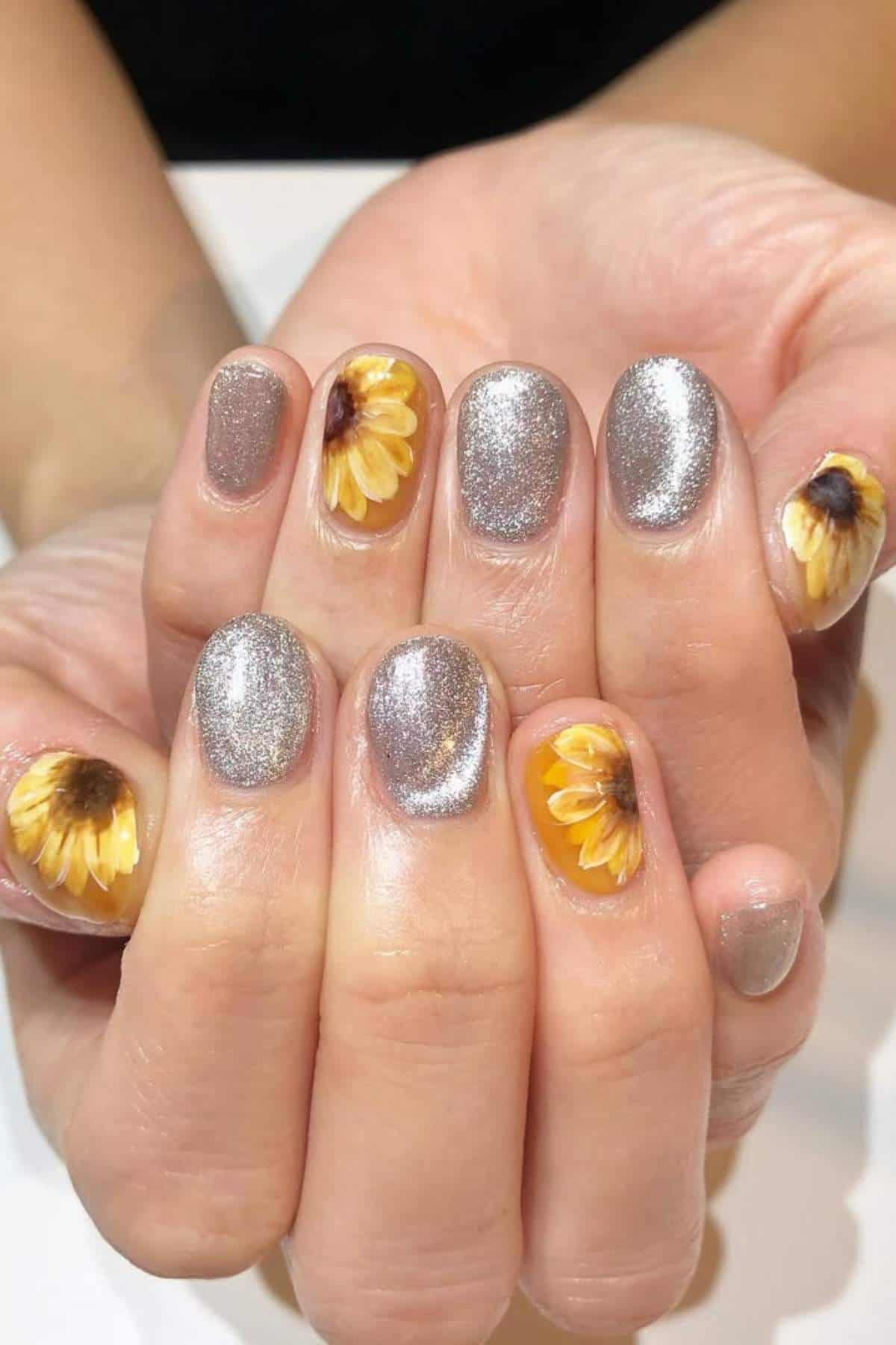 Hands with manicured nails; most nails painted metallic silver, while both thumb and pinky nails feature detailed sunflower designs.