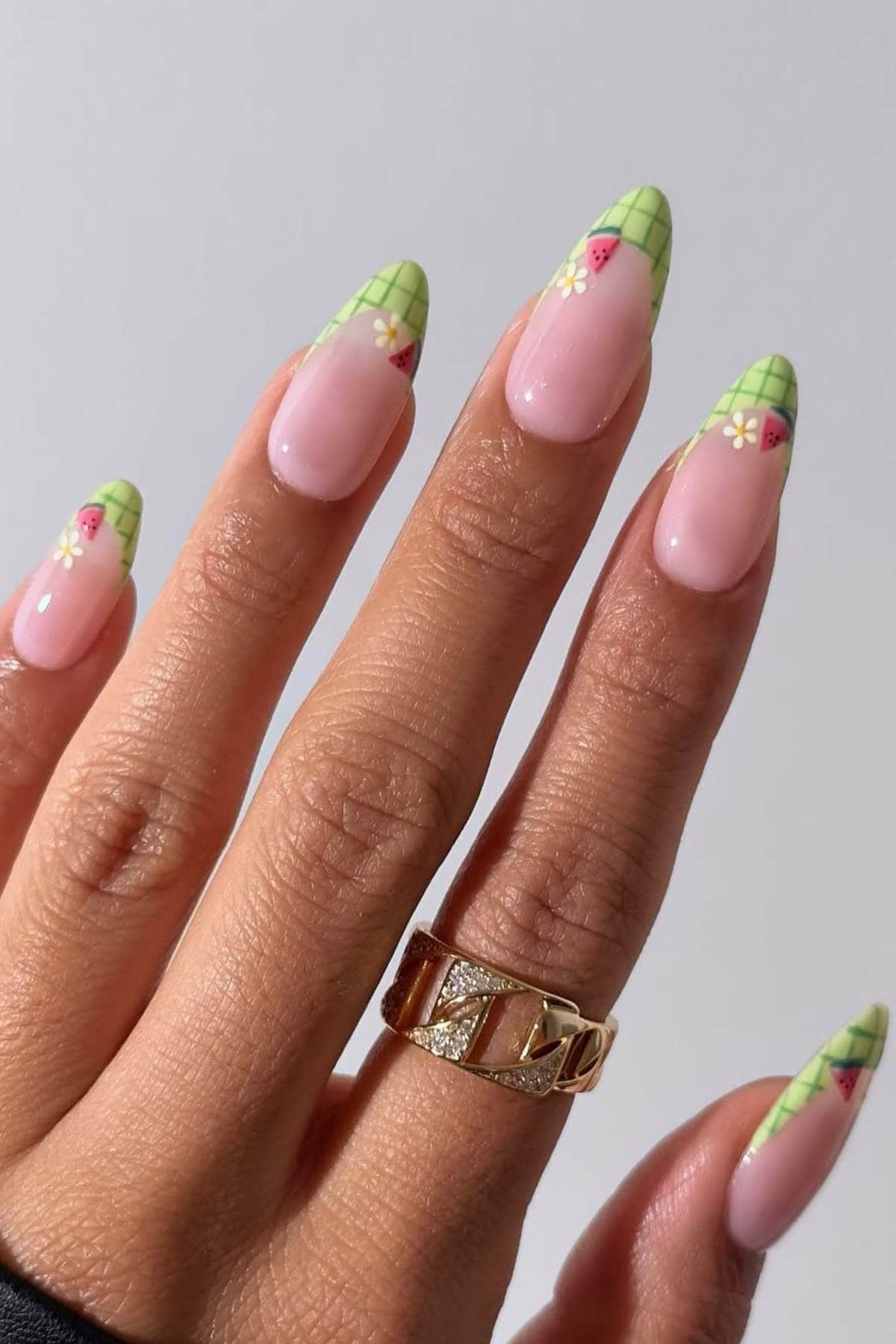 A hand with long almond-shaped nails featuring a green checkered French tip design with small flowers and strawberries. The person is wearing a gold ring.