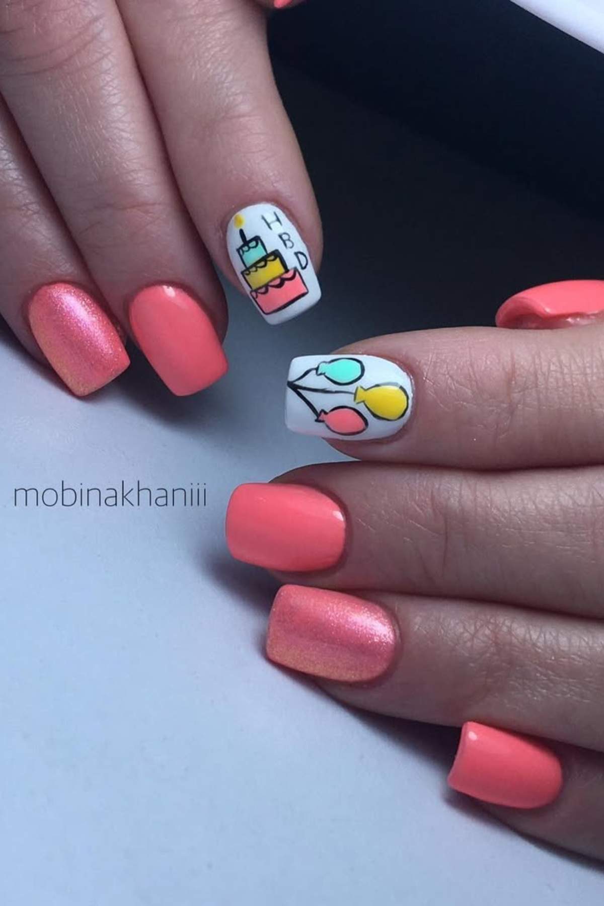 A hand with coral pink nails; one nail features a birthday cake and "HBD," and another has colorful balloon nail art.