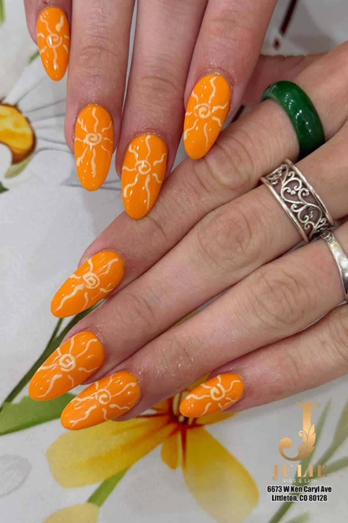 Hands with long, almond-shaped nails painted orange and decorated with white floral designs. The person is wearing silver and green rings. A salon logo is visible in the lower right corner.