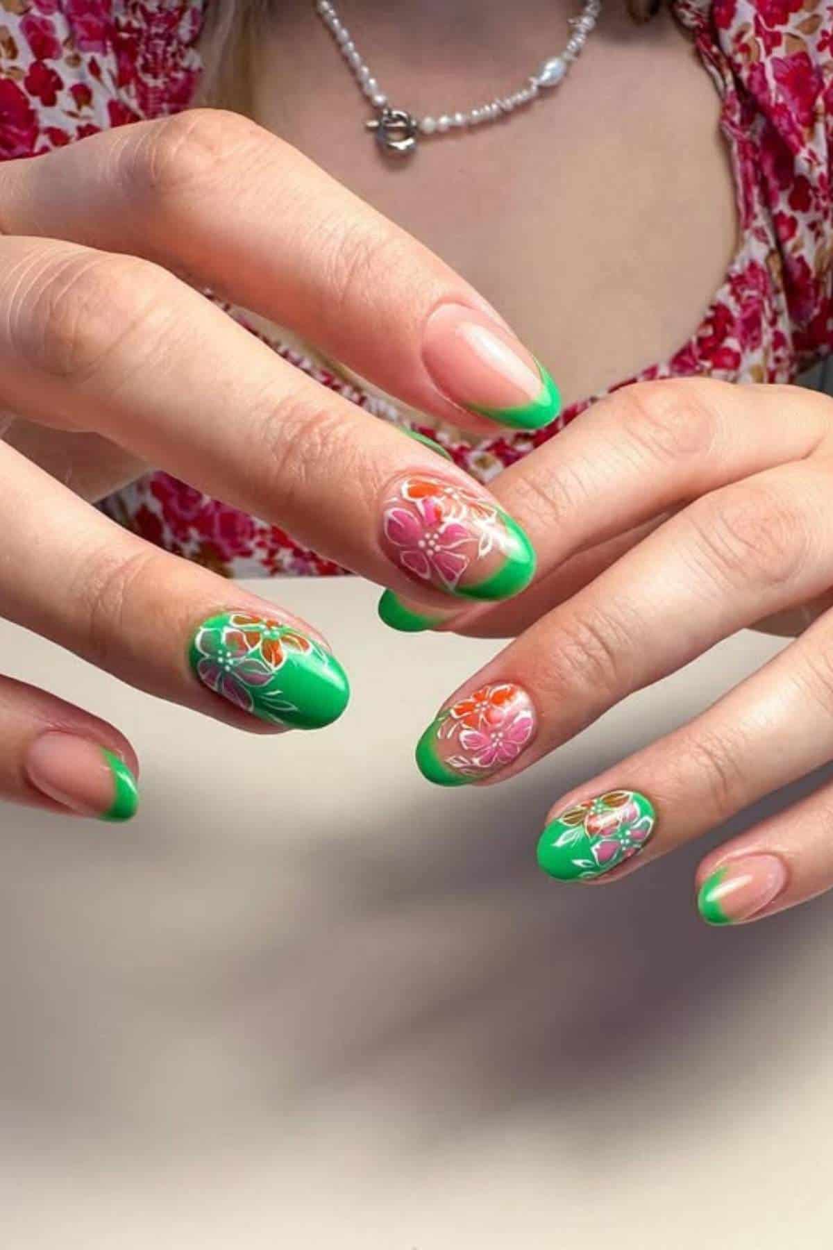 A person with green French tip nails featuring hand-painted floral designs in pink, orange, and white, wearing a pink patterned top and a beaded necklace.