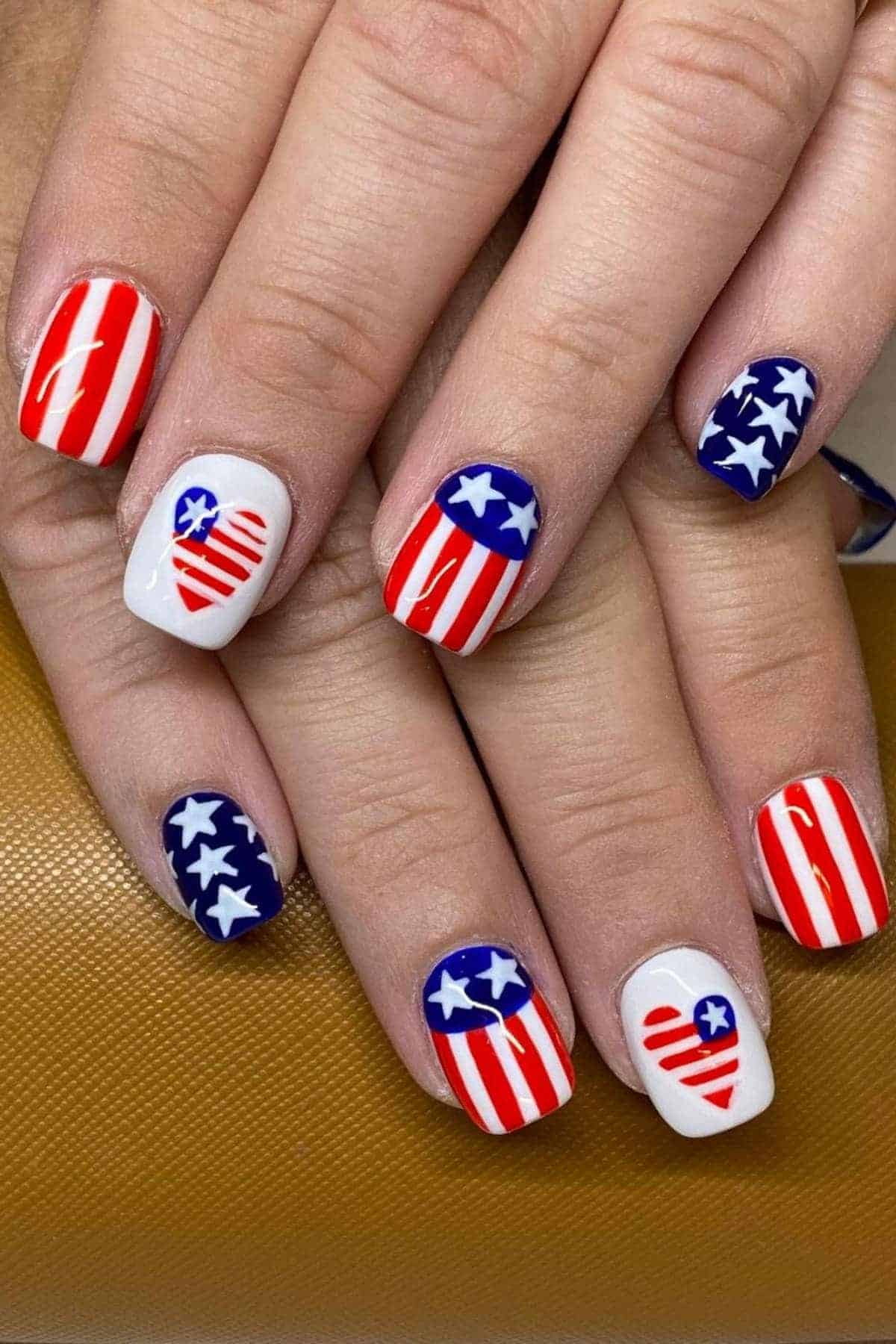 Close-up of hands with nails painted in red, white, and blue designs featuring stars and stripes, resembling the American flag.
