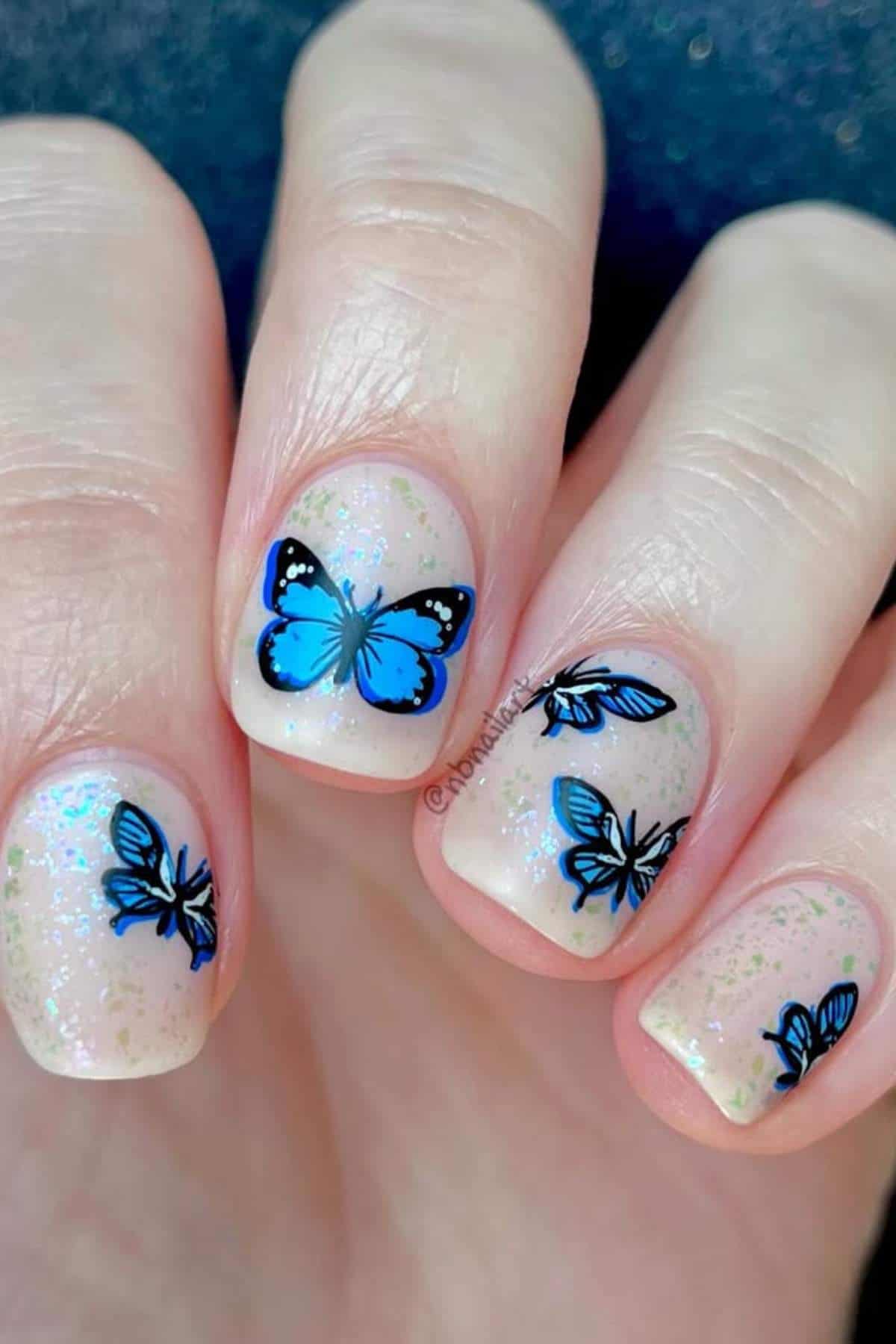 A close-up of fingernails painted with a light, shimmery polish and decorated with blue butterfly nail art designs.