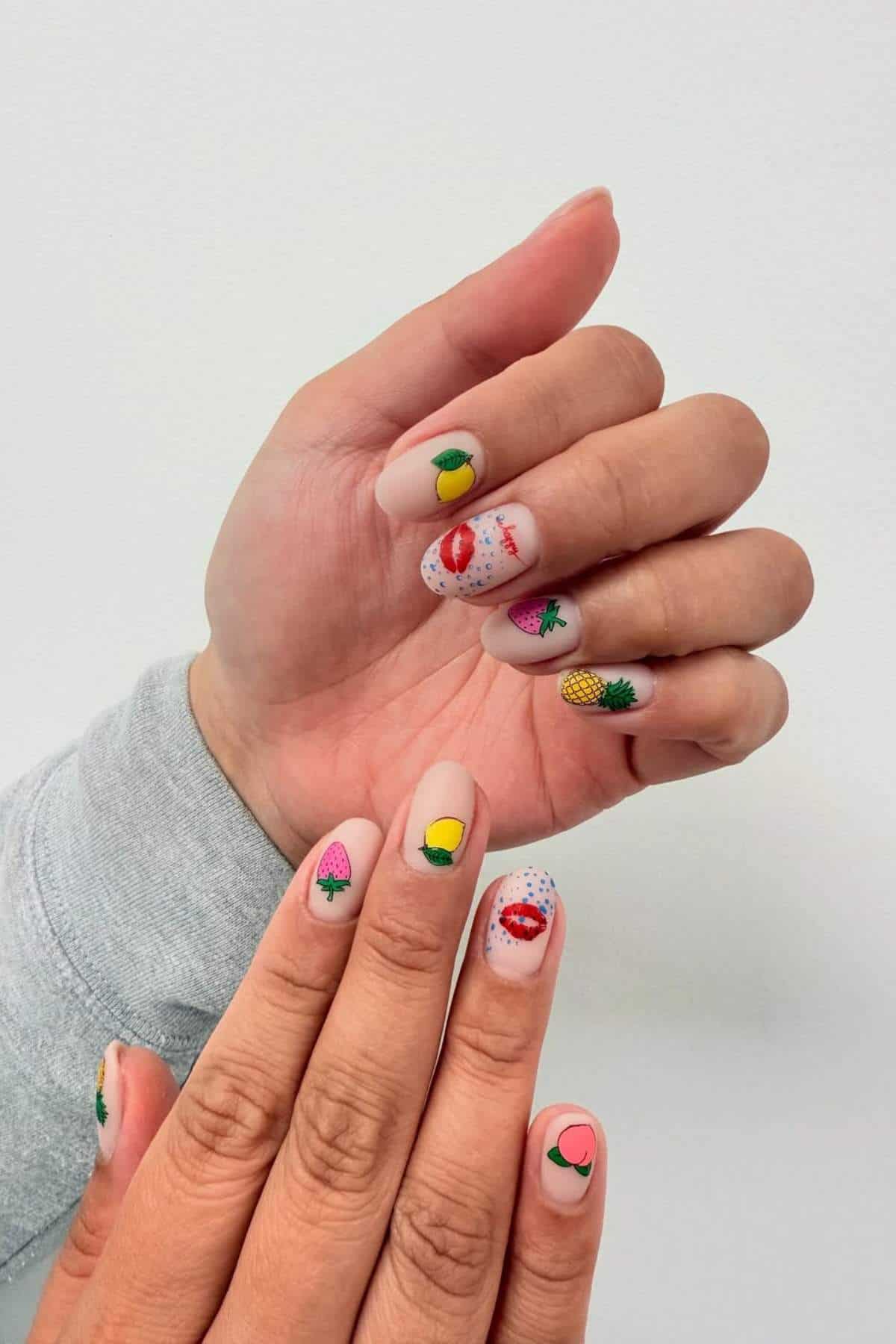 Two hands with light-colored nail polish featuring small, colorful fruit designs including strawberry, lemon, orange, pineapple, and kiwi on each fingernail.