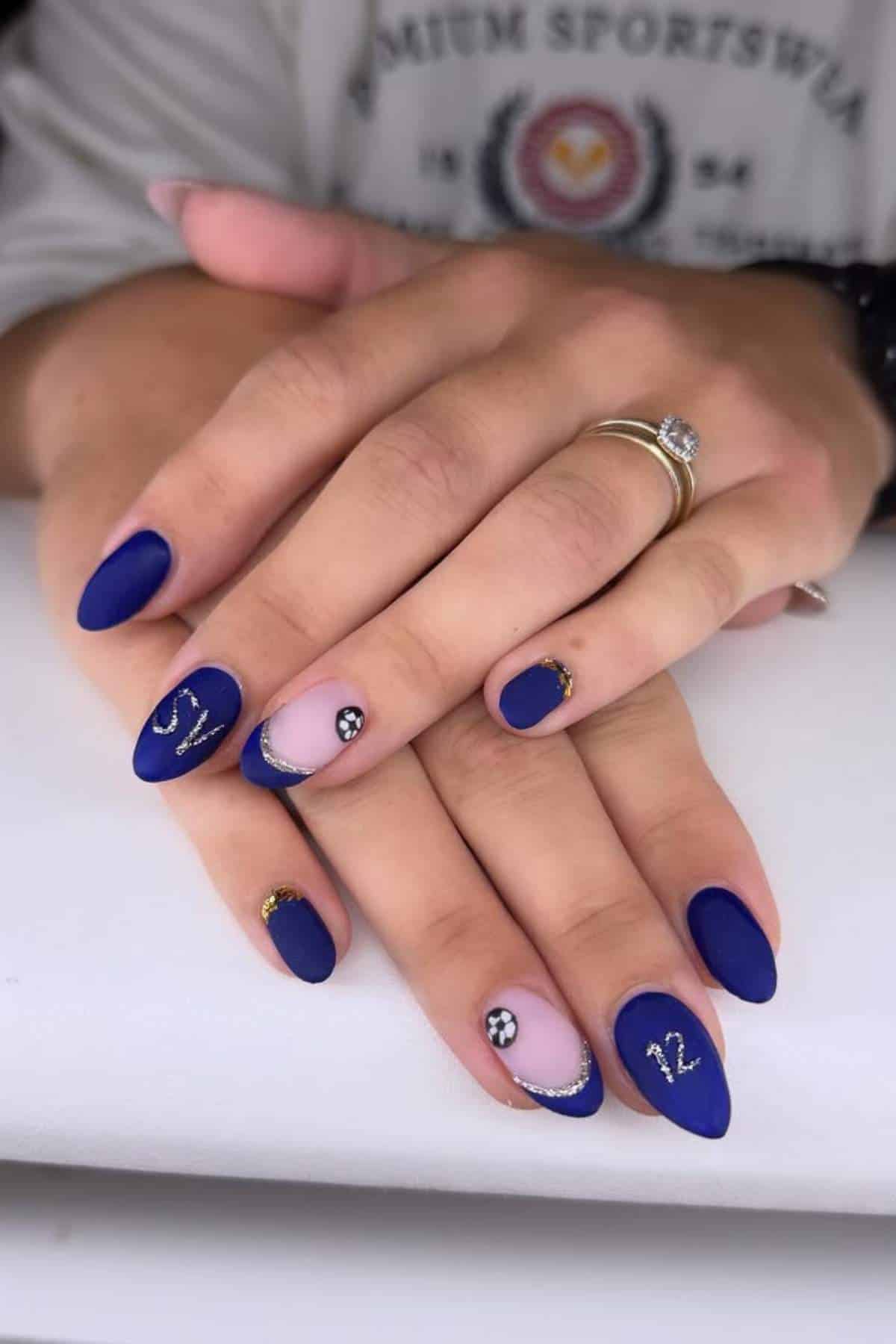 Hands with matte royal blue and nude almond-shaped nails, decorated with silver glitter, small rhinestones, and the numbers 12 and 17 on the ring and pinky fingers.