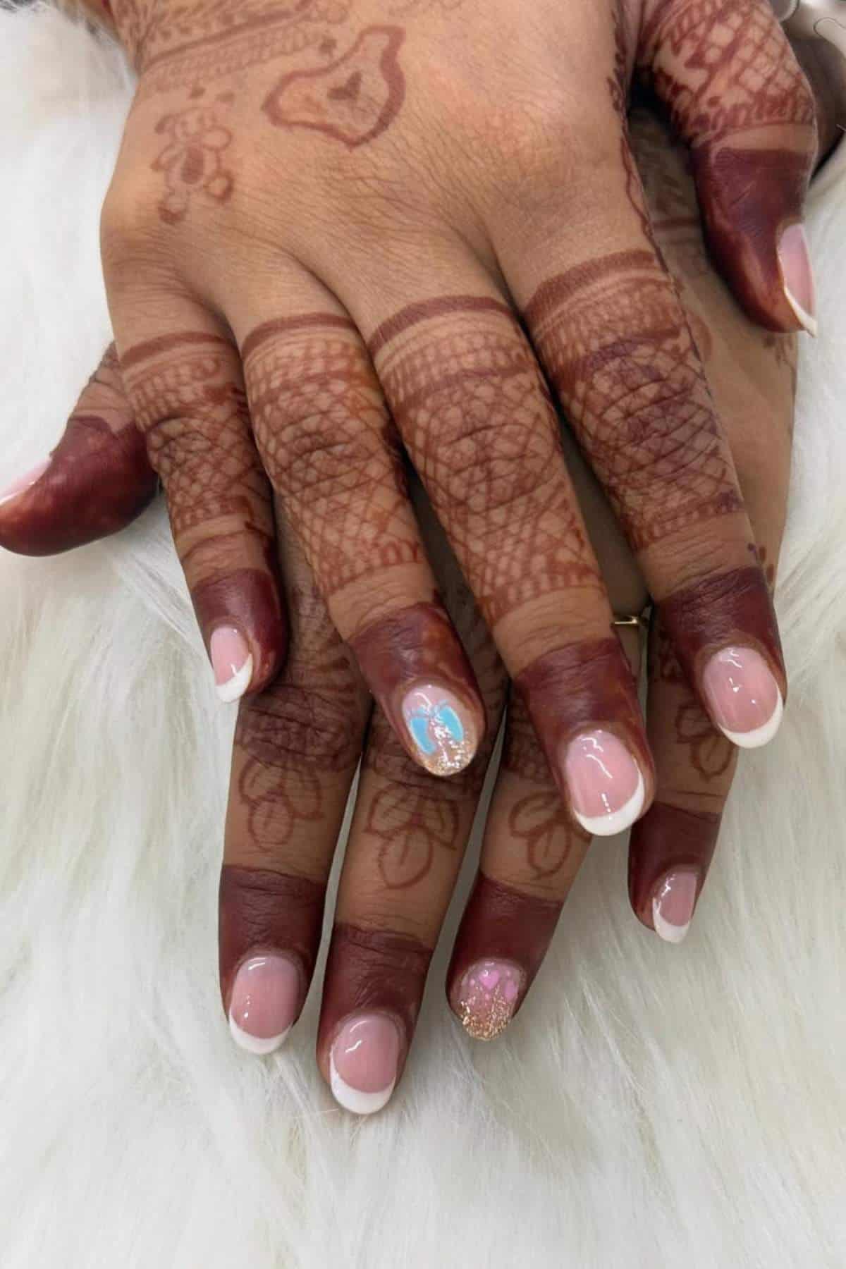 Two hands with intricate henna designs on the skin, displaying French manicured nails; one nail features blue and white nail art, and another has gold glitter.