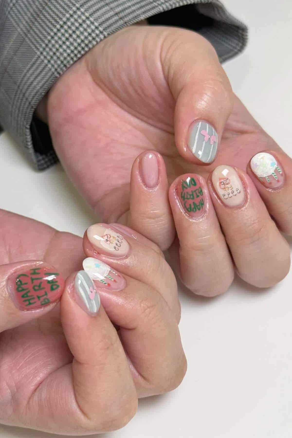 A close-up of hands with short nails featuring colorful birthday-themed nail art, including text, balloons, candles, and decorative designs.