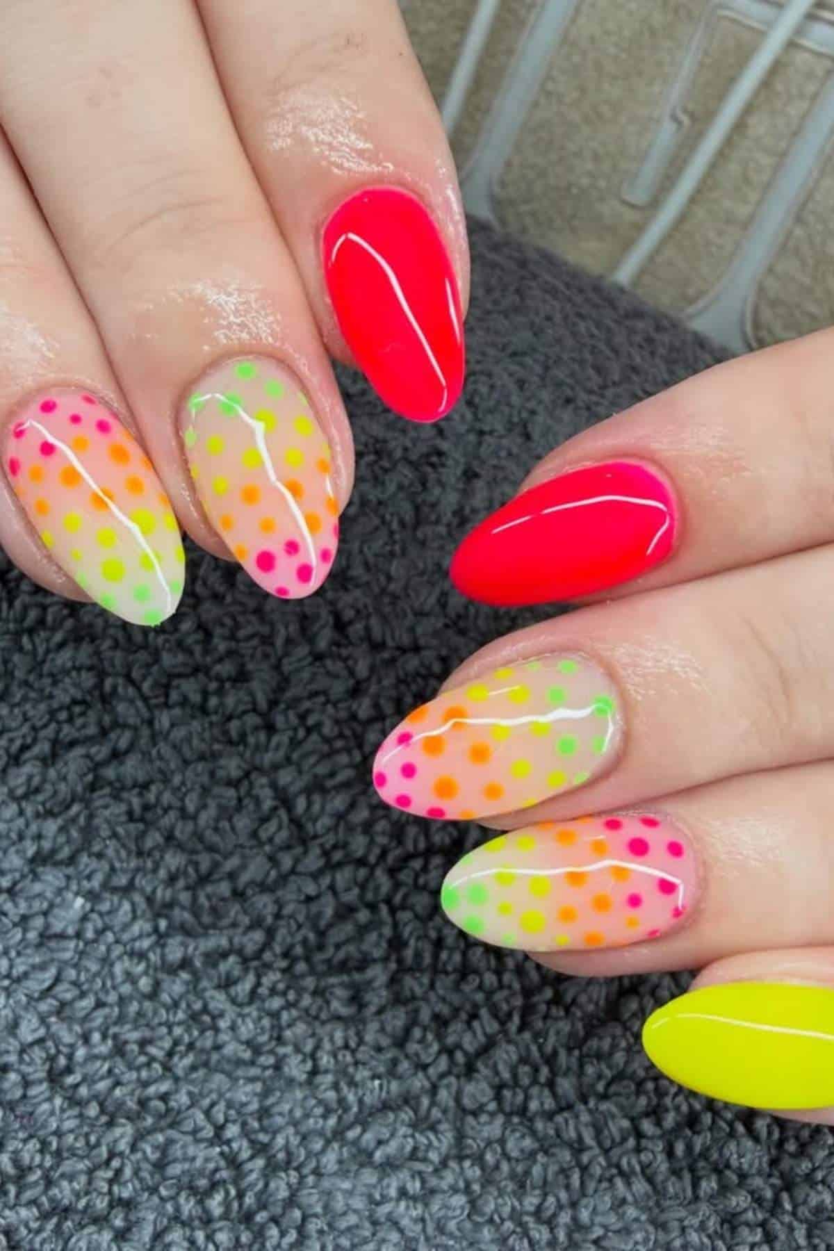 Hands with almond-shaped nails featuring neon colors and polka dot designs in pink, green, orange, and yellow on a dark textured surface.