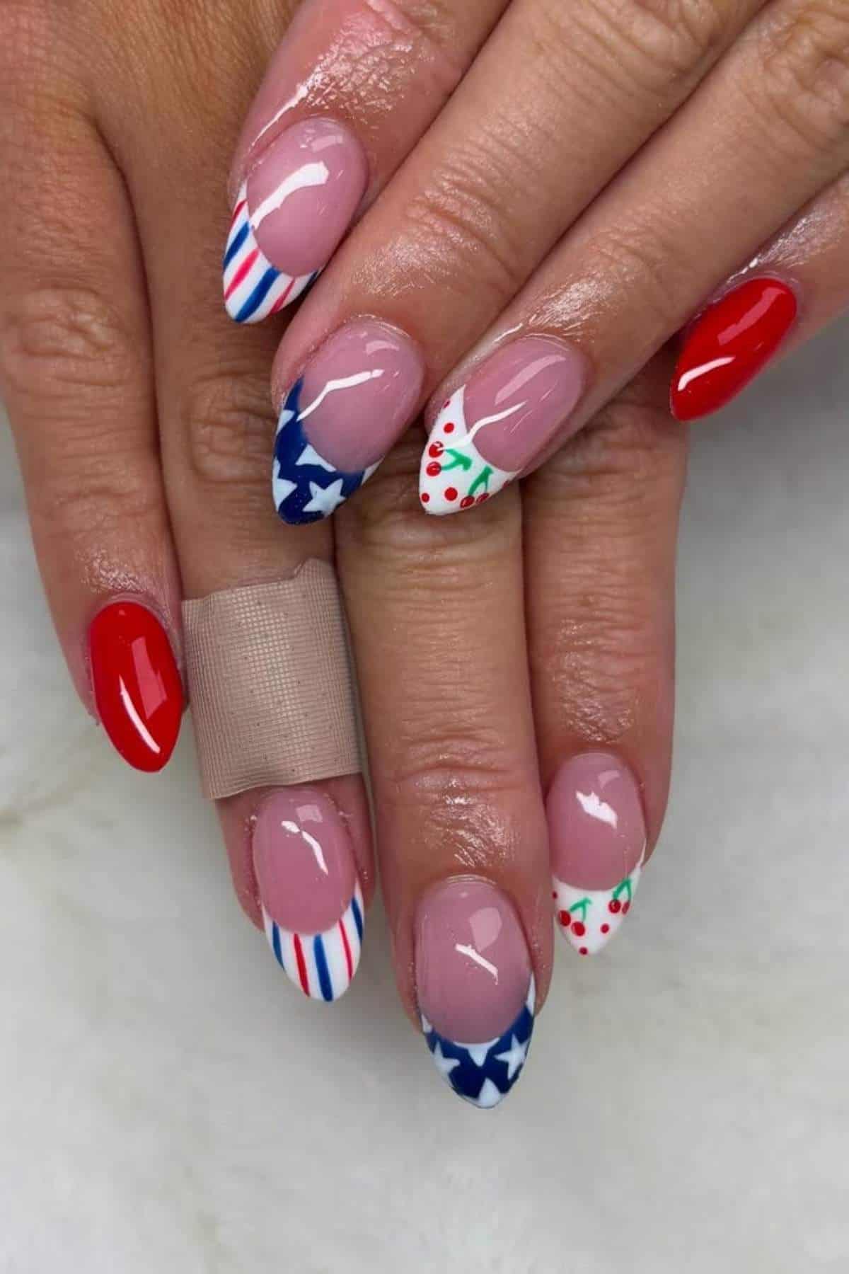 Hands with oval-shaped nails featuring red, white, and blue patriotic designs, including stars, stripes, cherries, and red polish; one finger has a bandage.