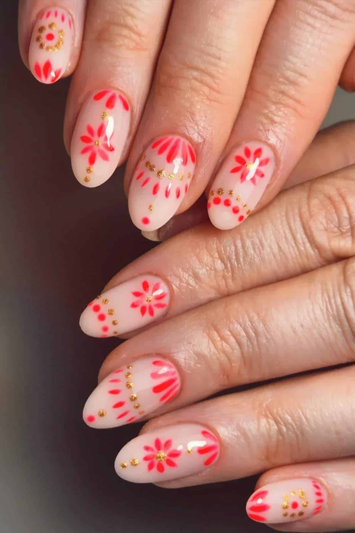 Hands with almond-shaped nails featuring a pink and coral floral design, gold dots, and nude background. Nails are neatly manicured and decorated with detailed patterns.