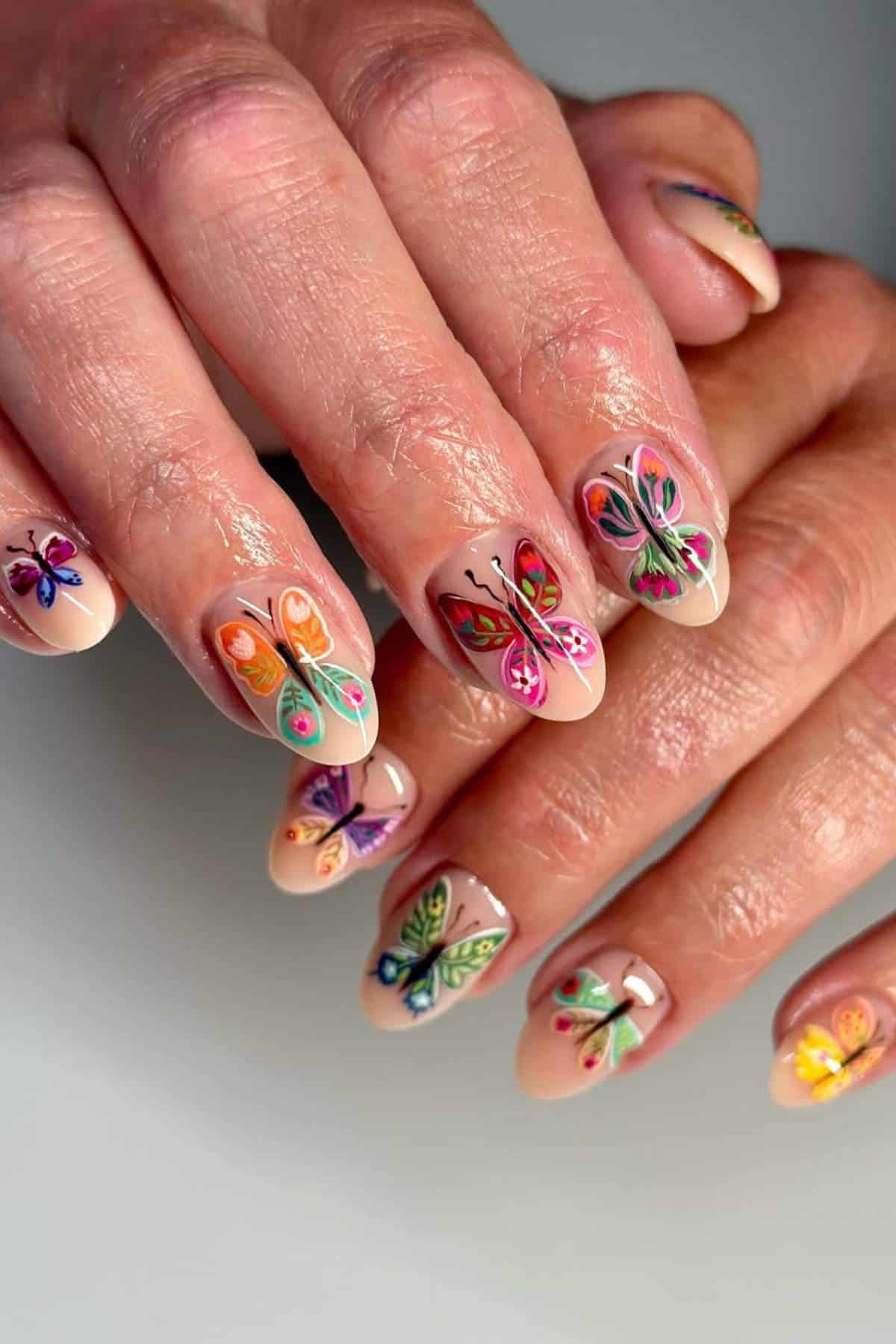 Close-up of hands with nude-colored nails featuring colorful butterfly nail art on each fingernail.