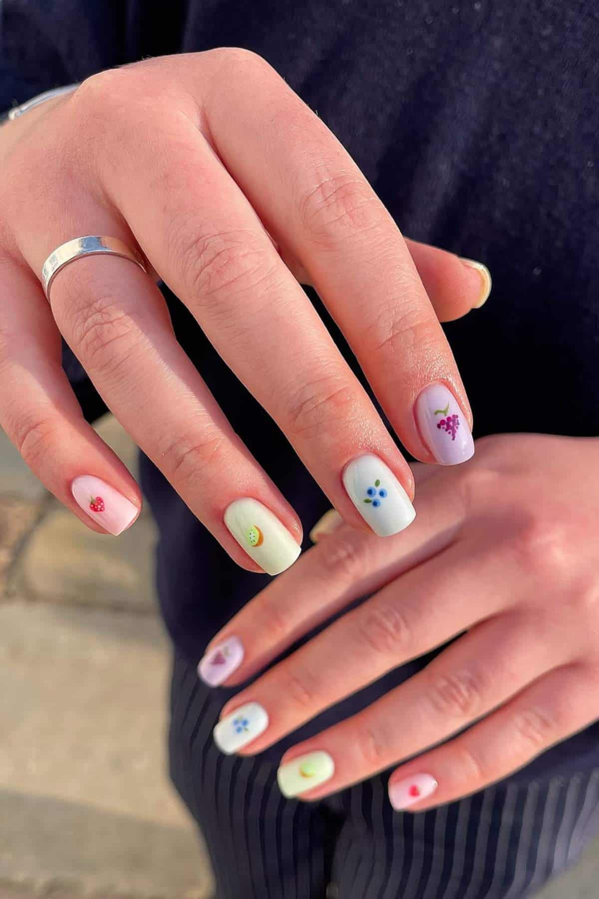 A person displays manicured nails painted in pastel colors with small, colorful fruit and flower designs on each nail.