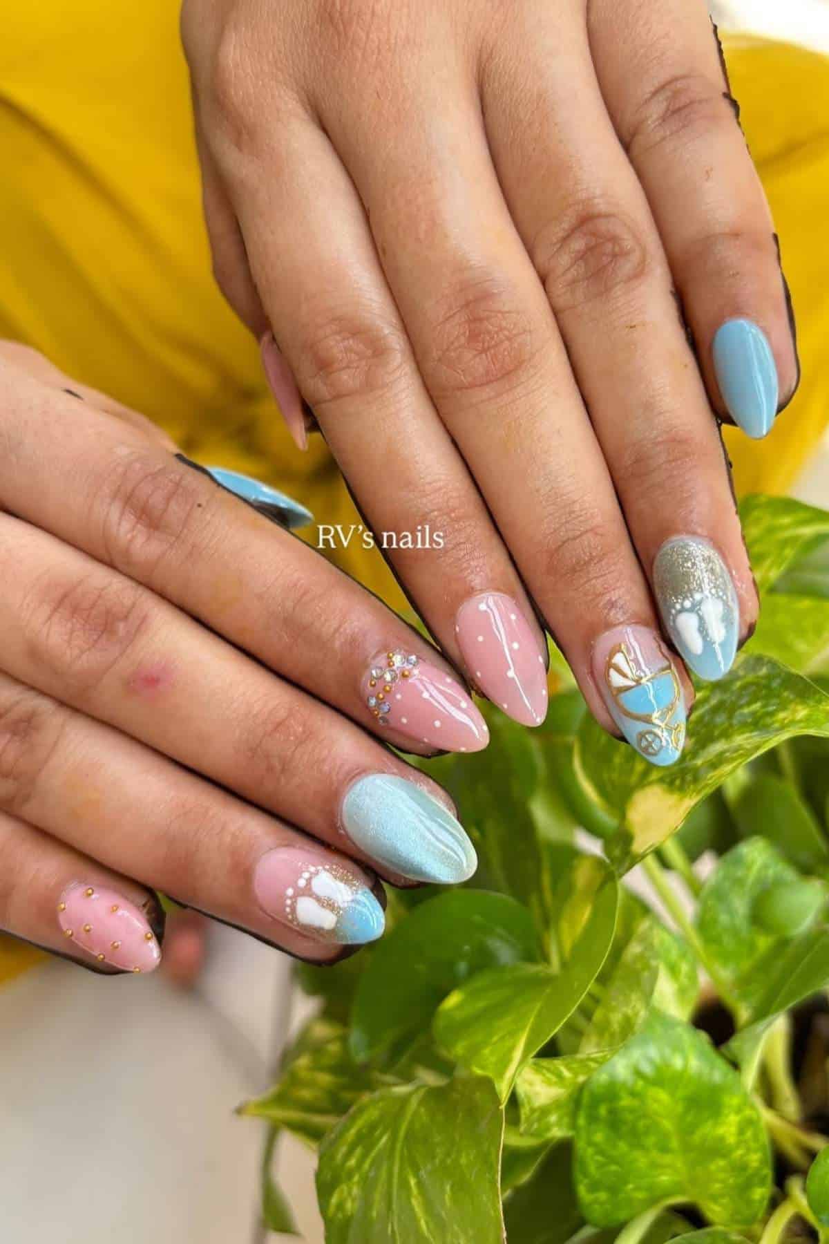 Two hands with almond-shaped nails painted in pastel blue and pink, featuring various designs, pearls, studs, and glitter accents, held above green leaves.