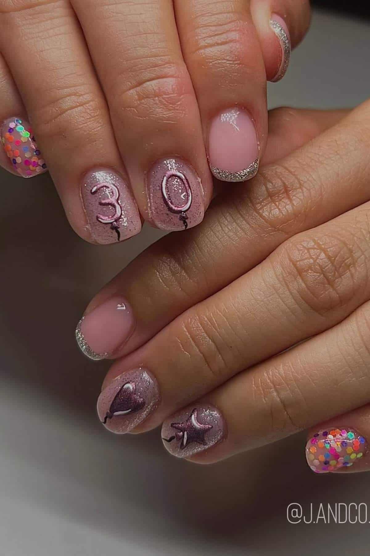A close-up of hands with manicured nails, featuring pink glitter polish, silver tips, and "30" with a balloon and star design; some nails display colorful confetti.