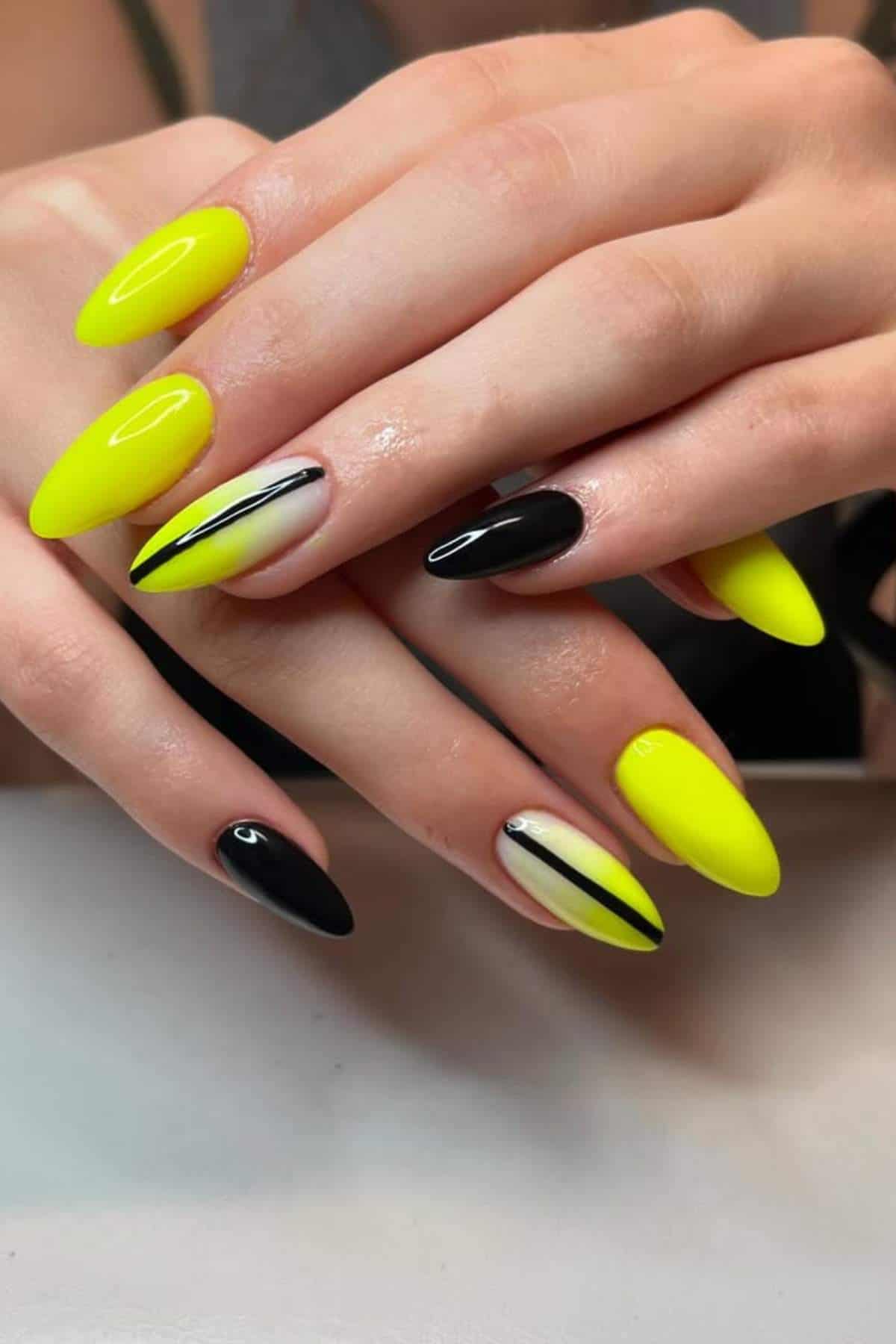 A hand with almond-shaped nails painted in yellow, black, and nude colors, featuring black and yellow diagonal stripe designs on two accent nails.