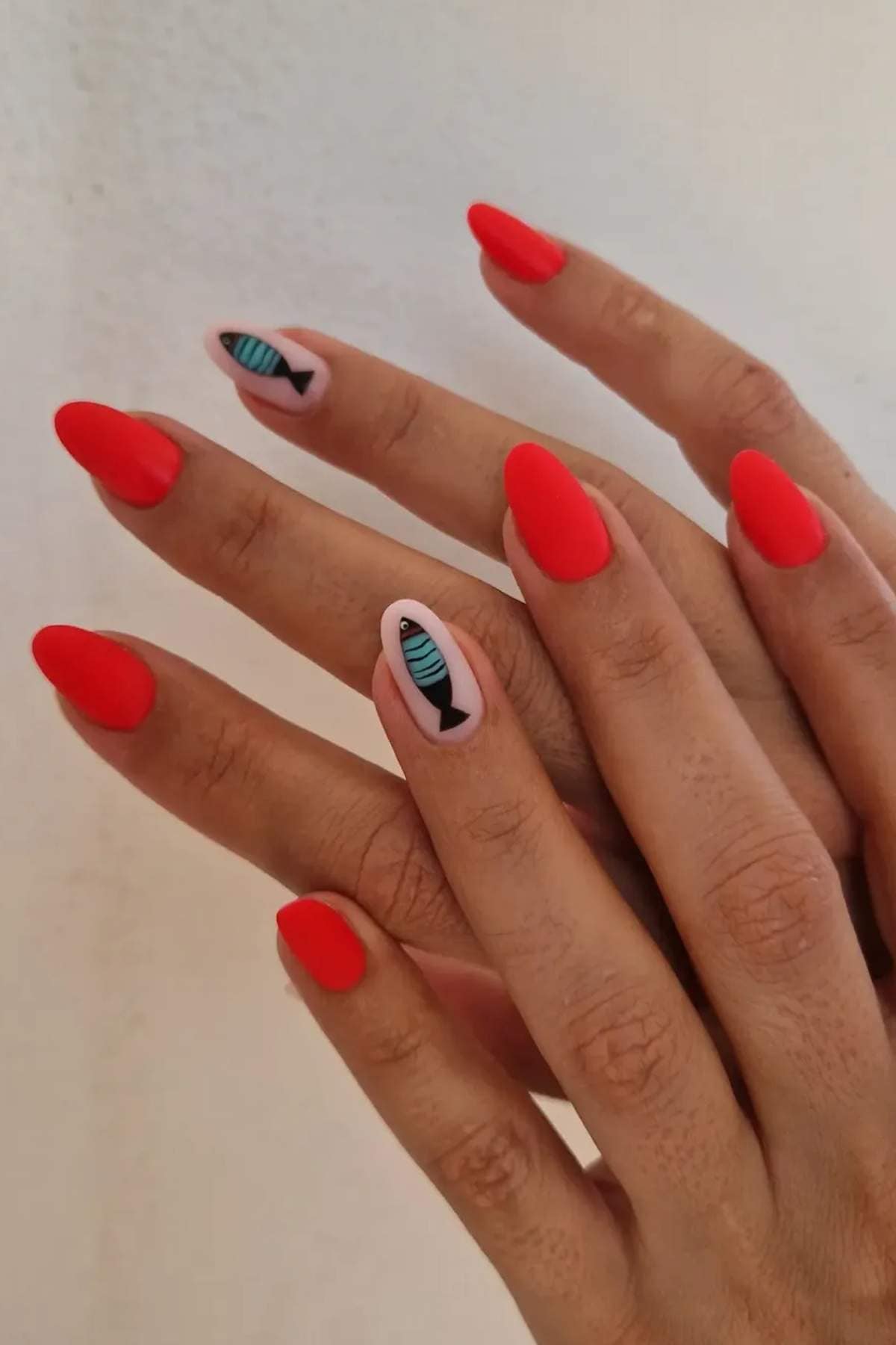 Hands with matte red almond-shaped nails; ring fingers feature nail art of a black fish outline with a blue striped pattern on a light pink background.