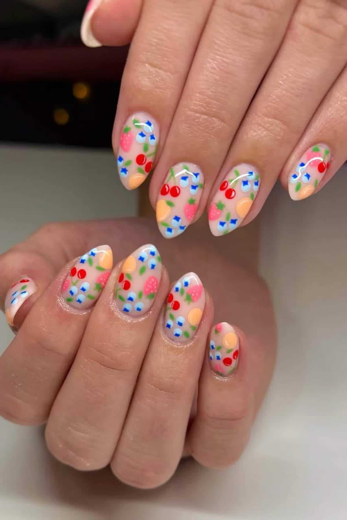 Close-up of hands with almond-shaped nails painted with colorful fruit designs, including cherries, strawberries, and blueberries, on a nude base.