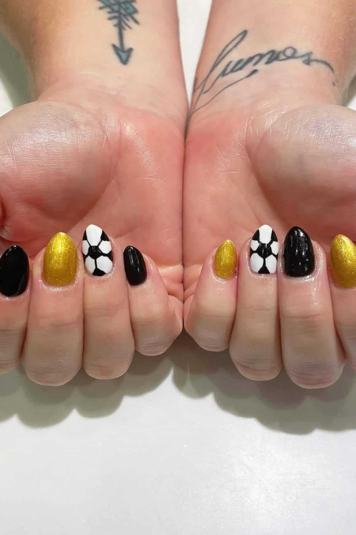 Two hands display manicured nails painted in black, gold, and white, with a soccer ball pattern on two nails. Black tattoos are visible on the wrists.