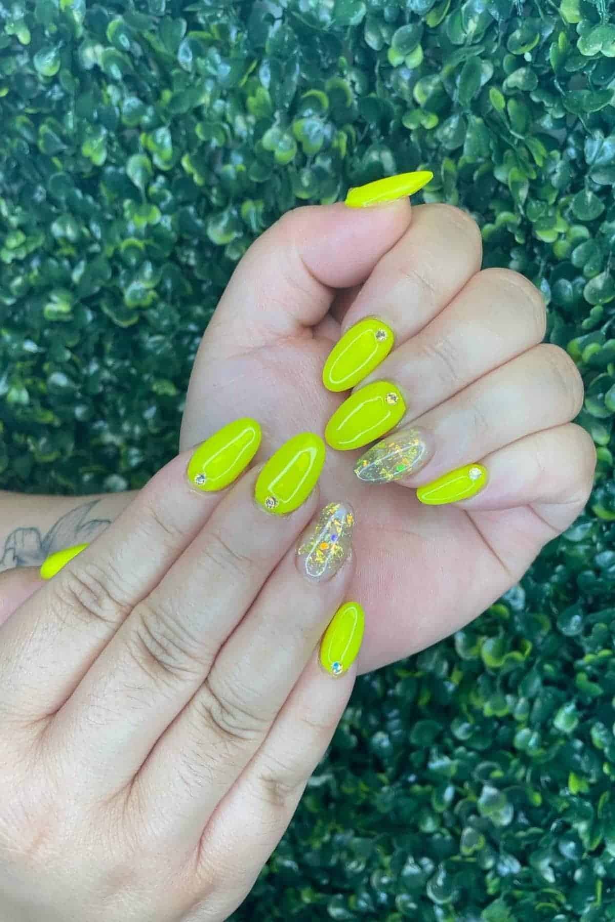 Hands with bright yellow oval-shaped manicured nails, some with small rhinestones and two accent nails featuring clear tips with iridescent foil. Green leafy background.